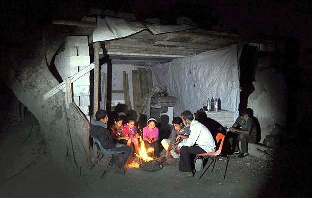 L'image montre un groupe de personnes assises autour d'un feu dans un environnement sombre. Ils semblent rassemblés à l'intérieur d'une structure rudimentaire, faite de matériaux simples comme du bois et des tôles. Les gens, probablement des adultes et des enfants, sont concentrés sur le feu, qui éclaire leur visage et crée une atmosphère chaleureuse malgré les conditions environnantes. Des éléments comme des bouteilles sont visibles en arrière-plan, suggérant une ambiance de partage et de convivialité, malgré le manque de confort.