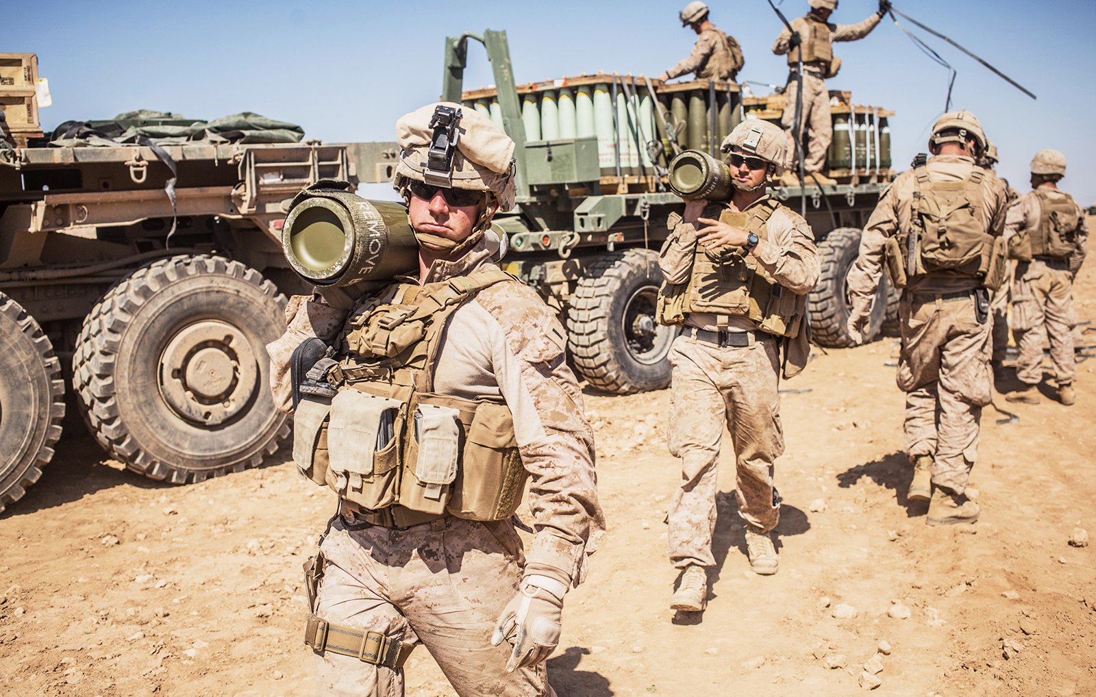 L'image montre des soldats en tenue militaire, probablement en opération. Certains d'entre eux portent des gilets tactiques et des casques, tandis qu'un soldat en avant tient un obus de mortier. En arrière-plan, on aperçoit un véhicule militaire avec une remorque et d'autres soldats s'affairant. Le cadre semble désertique, avec un sol sablonneux et un ciel dégagé. On ressent un climat de préparation et d'activité militaire.