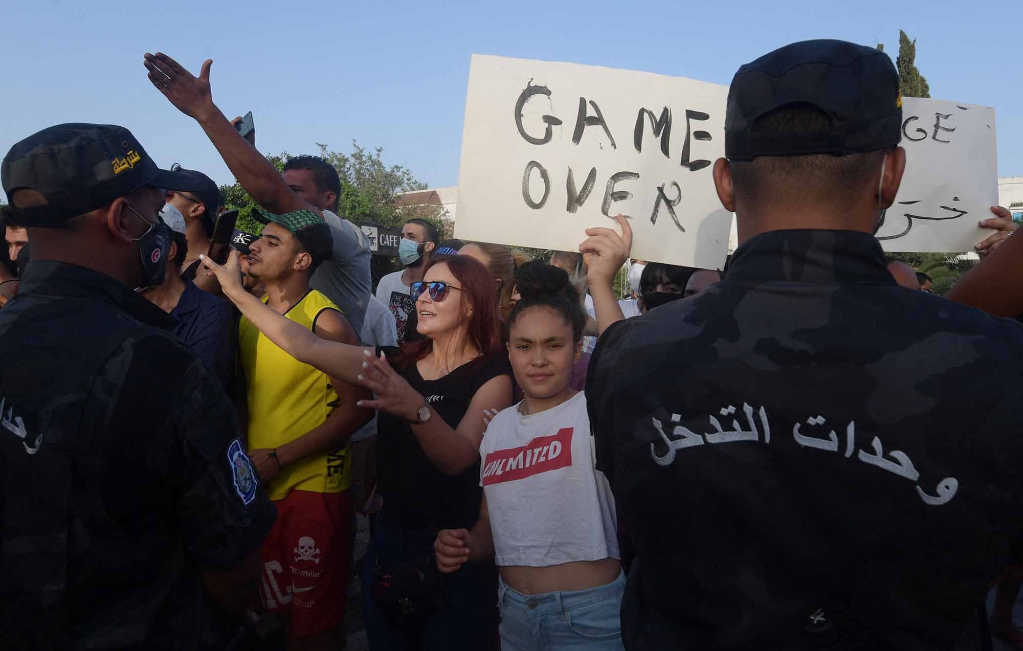 Dans l'image, on peut voir une scène de manifestation avec un groupe de personnes se rassemblant. Certains manifestants tiennent des pancartes, dont l'une indique "GAME OVER". Des policiers en uniforme, faisant face aux manifestants, semblent surveiller la situation. L'ambiance est tendue, avec des expressions variées sur les visages des participants, certains montrant de la détermination. L'arrière-plan suggère un lieu public, probablement en milieu urbain.