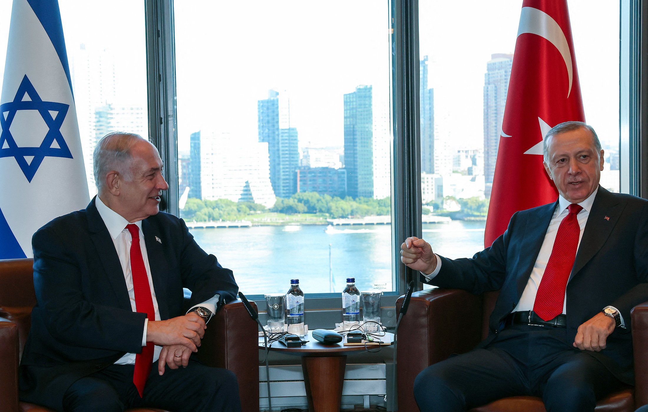 L'image montre deux hommes en discussion dans un cadre professionnel. À gauche, il y a un homme avec une cravate rouge, assis sur une chaise, regardant l'autre homme. À droite, l'autre homme, également en cravate, semble parler et gesticuler. En arrière-plan, on aperçoit des gratte-ciel et une vue sur l'eau, suggérant un environnement urbain. Des drapeaux d'Israël et de Turquie sont visibles, indiquant le contexte de cette rencontre.