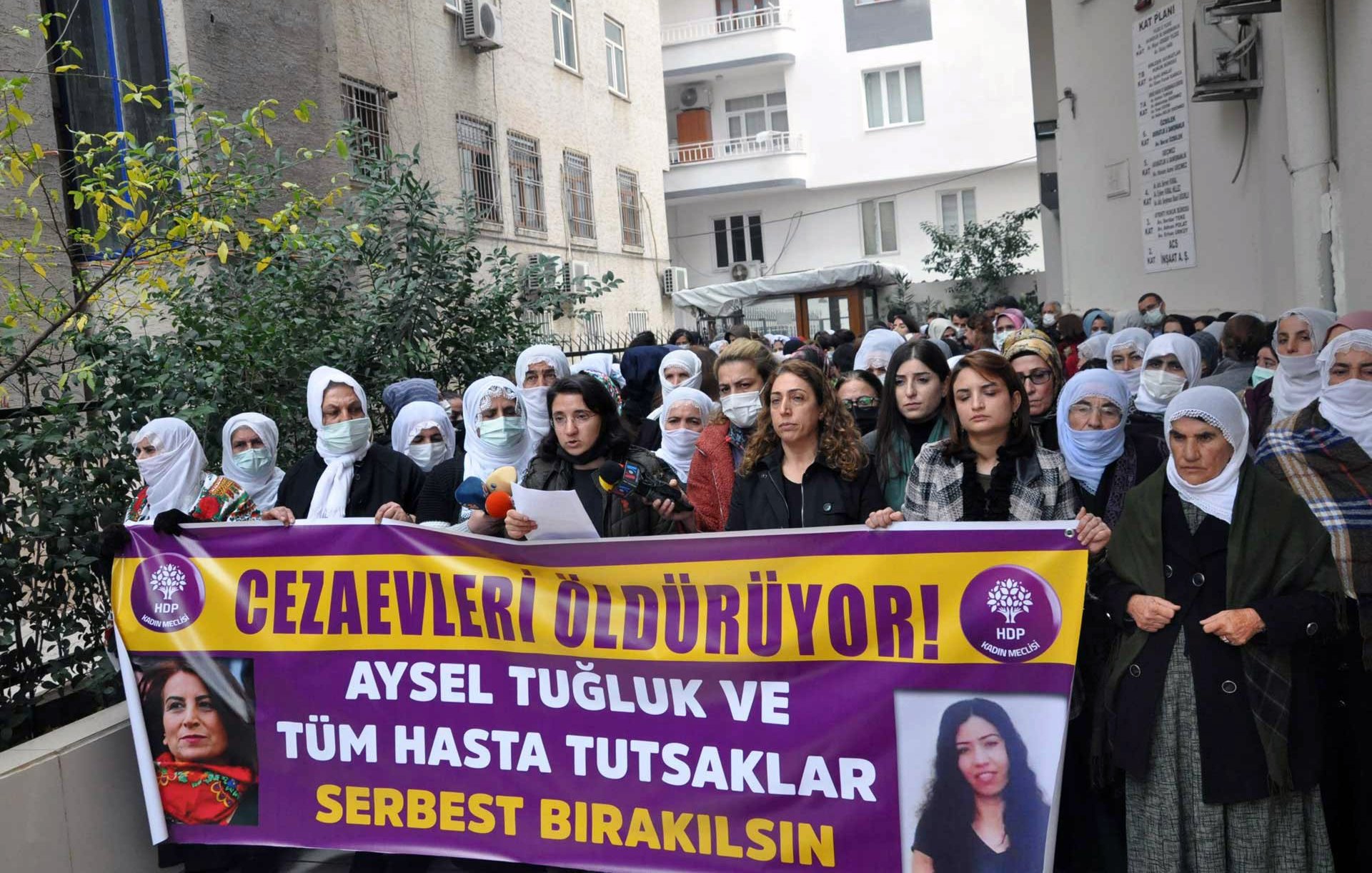 L'image montre un groupe de personnes rassemblées dans une rue, tenant des pancartes et des banderoles. Les femmes portent des foulards sur la tête et semblent soutenir une cause ou un mouvement. L'une des banderoles porte des slogans demandant la libération d'Aysel Tuğluk et des prisonniers malades. Le contexte semble être une manifestation pour les droits de l'homme et la justice. Des expressions de détermination et de solidarité sont visibles parmi les participants.