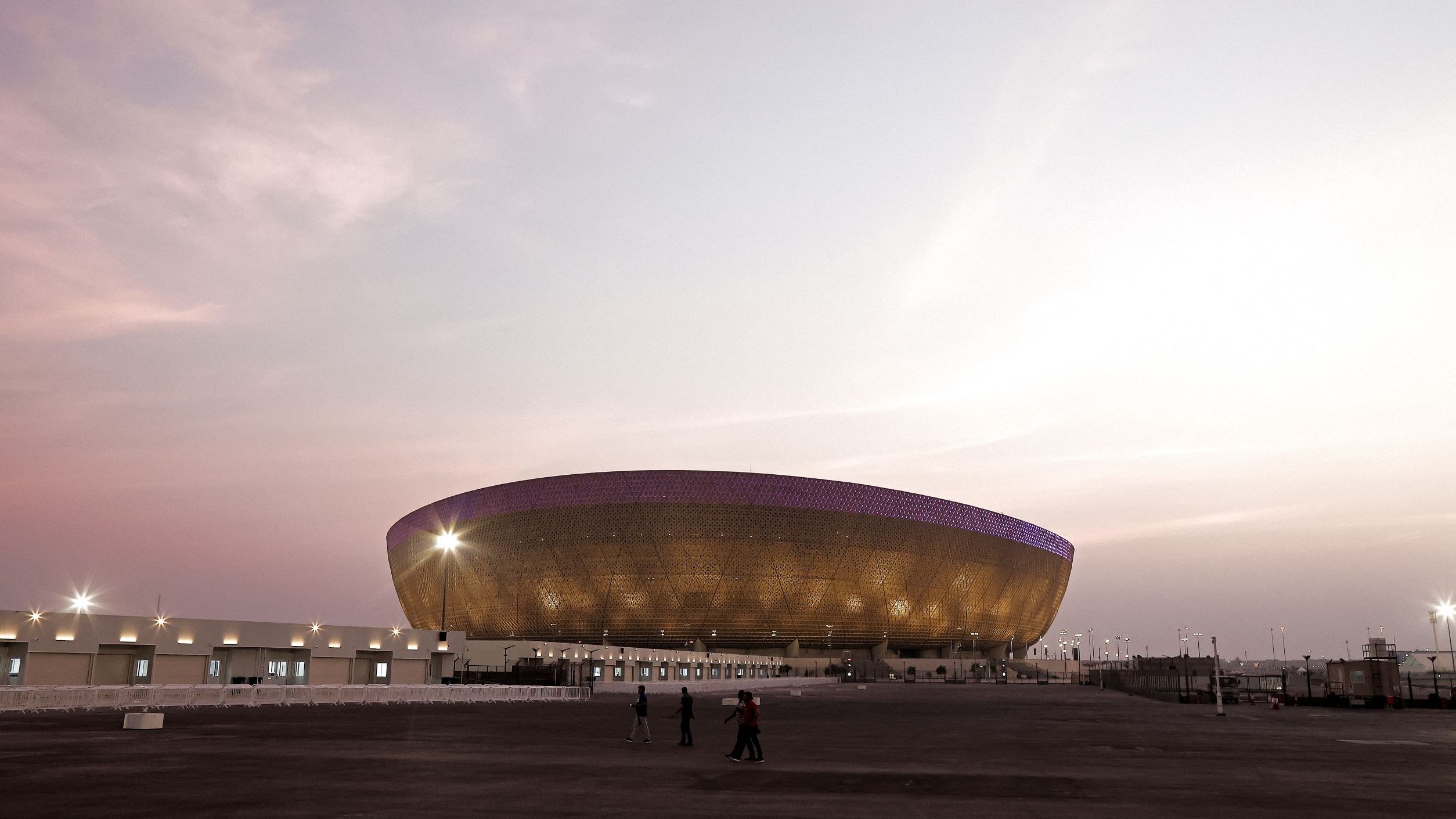 L'image montre un grand stade moderne à l'architecture distinctive, situé sous un ciel teinté de nuances pastel au coucher du soleil. Le stade possède une structure en forme d'ellipse avec une façade brillante, peut-être en métal ou en verre, qui capte la lumière ambiante. On aperçoit quelques personnes marchant devant le stade, soulignant sa taille imposante. L'ensemble de la scène dégage une ambiance calme et majestueuse.