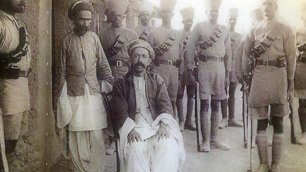 L'image montre un groupe de personnes en tenue traditionnelle, avec un homme assis au premier plan, semble-t-il en position plus détendue. Il porte des vêtements amples et un turban. Autour de lui, plusieurs hommes en uniforme militaire se tiennent debout, équipés de ceintures et de fusils, donnant une impression de formation ou de cérémonie. L'environnement semble être un lieu extérieur, probablement historique.
