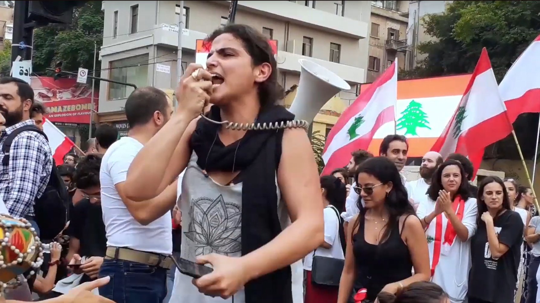 Foule en manifestation, femme au mégaphone, drapeaux libanais en arrière-plan.