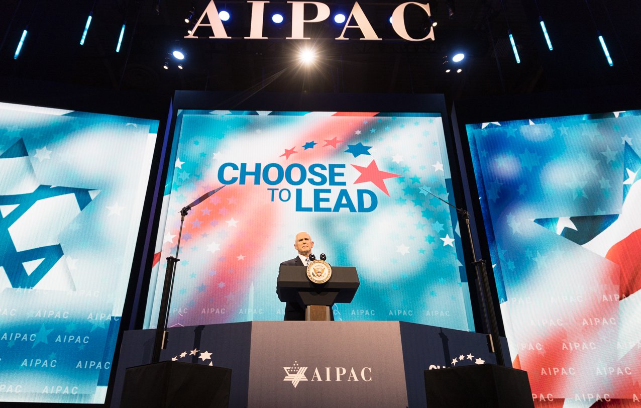 L'image montre un homme sur scène, probablement un orateur, devant un public. Il se tient derrière un pupitre avec le logo d'AIPAC, une organisation pro-israélienne. En arrière-plan, il y a un grand écran affichant le slogan "CHOOSE TO LEAD" (Choisissez de diriger), entouré de motifs étoilés. L'ambiance semble formelle et dédiée à un discours important, probablement lors d'une conférence ou d'un événement lié à AIPAC.