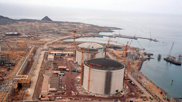 L'image montre un site industriel en construction, probablement une installation de gaz naturel ou une centrale électrique. On peut voir plusieurs grands réservoirs cylindriques, entourés de grues et de matériaux de construction. L'environnement est côtier, avec une vue sur la mer et des montagnes en arrière-plan. Il y a également des routes et des véhicules de construction présents sur le site. Le paysage est principalement désertique et montagneux.