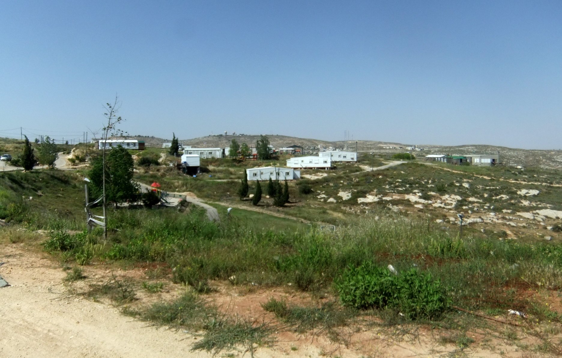 L'image montre un paysage rural avec des collines à l'arrière-plan. On peut voir plusieurs maisons en métal ou modulaires, disposées de manière dispersée. La végétation est sparse, avec des zones de terre et de l'herbe verte. Le ciel est clair et ensoleillé. L'ensemble donne une impression d'isolement et de simplicité.