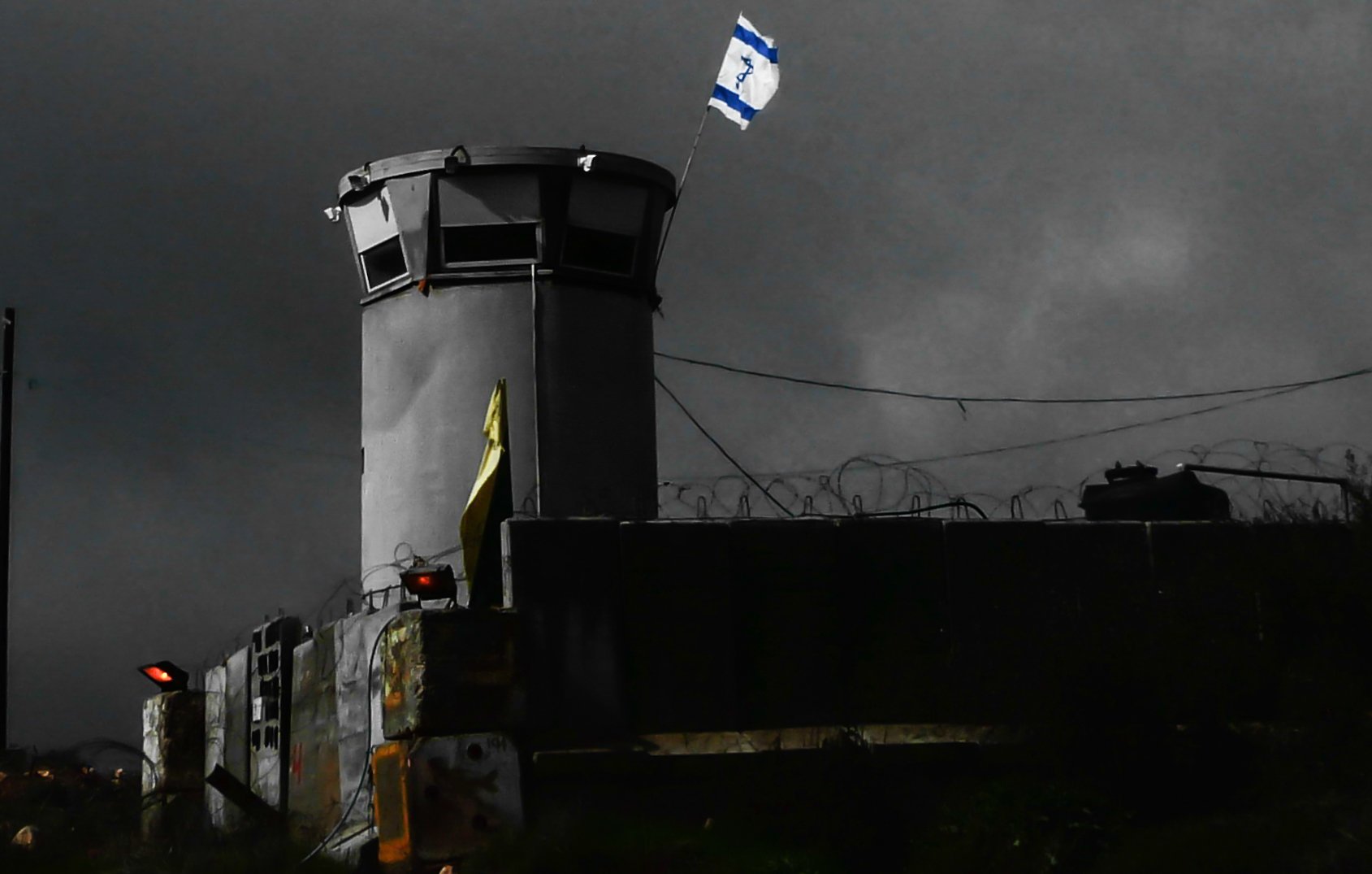 L'image montre une structure semblable à une torre de surveillance, située dans un environnement sombre et nuageux. On peut voir un drapeau israélien flottant au sommet de la tour. Des barbelés sont visibles autour du périmètre, ce qui suggère une zone sécurisée ou protégée. L'éclairage de la scène crée une atmosphère de tension, accentuant l'aspect austère du lieu.