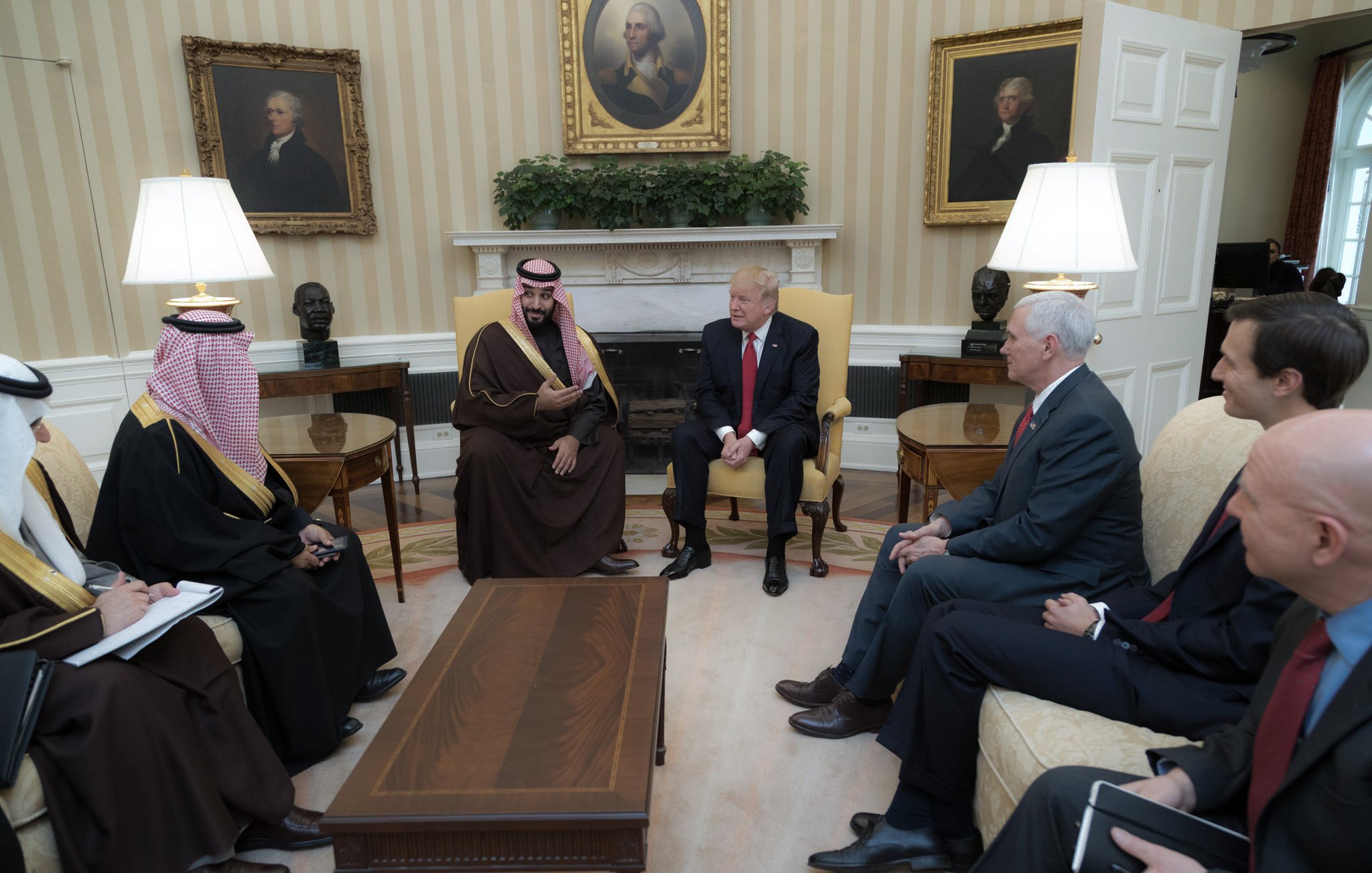 L'image montre une réunion dans un bureau d'un bâtiment officiel, probablement la Maison Blanche. On y voit plusieurs hommes assis autour d'une table basse. Au centre, un homme en costume rouge et cravate, vraisemblablement un dirigeant, discute avec un autre homme en tenue traditionnelle, qui porte un keffieh. D'autres personnes, habillées en costumes, sont également présentes, écoutant attentivement. En arrière-plan, on peut voir des portraits accrochés aux murs et des plantes décoratives. L'ambiance semble formelle et sérieuse, suggérant une discussion diplomatique ou politique.