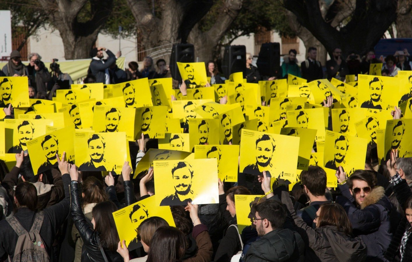 L'image montre une grande foule rassemblée, tenant des pancartes jaunes avec un visage représenté en noir. Les participants semblent mobilisés pour une cause, se tenant la main levée en signe de protestation ou de soutien. En arrière-plan, on peut voir d'autres personnes et des arbres, suggérant que l'événement a lieu en plein air. L'atmosphère paraît engagée et déterminée.