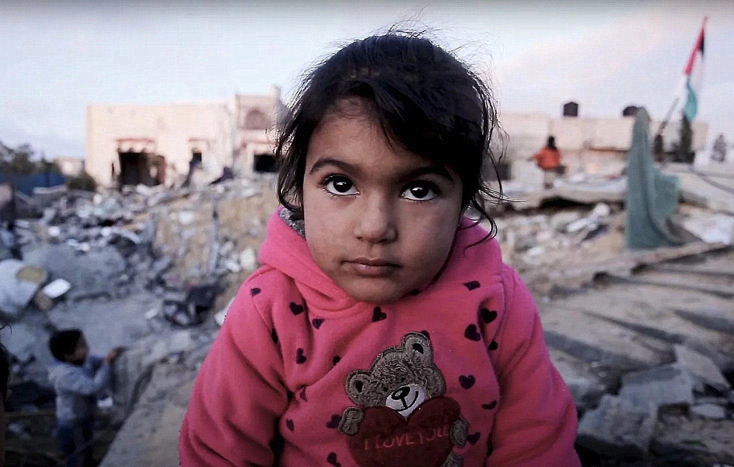 L'image montre une petite fille avec un regard expressif, vêtue d'un sweat à capuche rose orné d'un ours. Elle se trouve dans un environnement dévasté, entourée de décombres et de bâtiments en ruines. En arrière-plan, on aperçoit d'autres enfants. L'atmosphère semble chargée d'une grande tristesse et d'une vulnérabilité palpable.