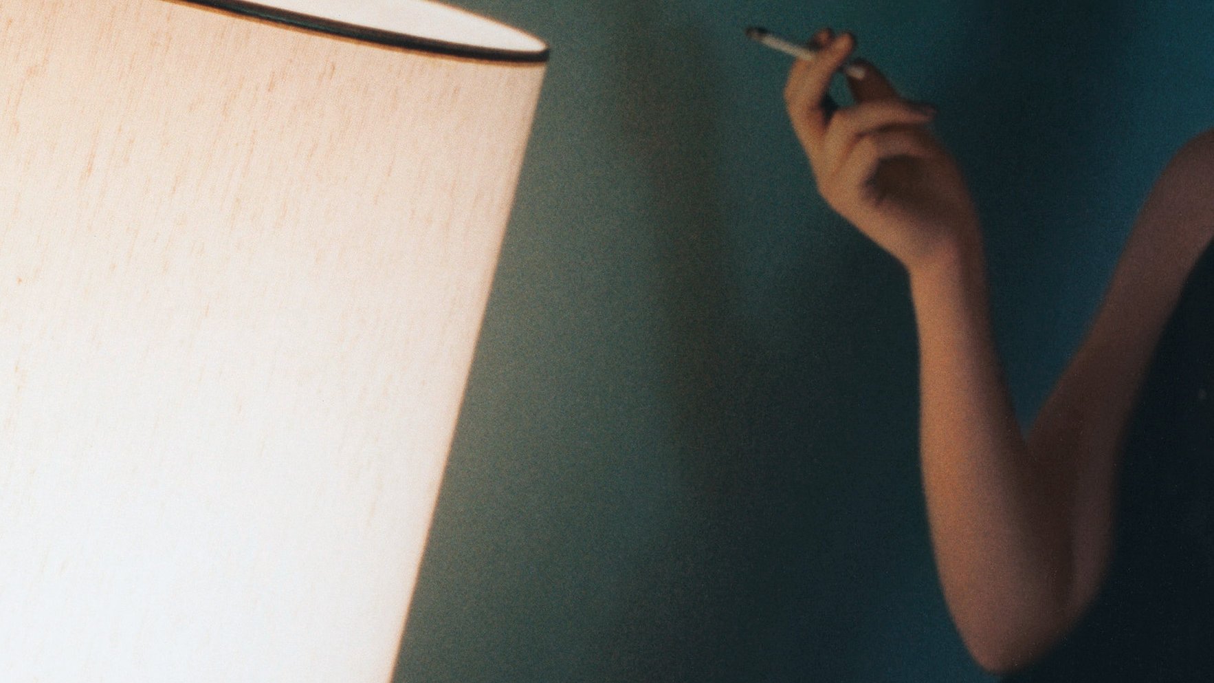 L'image montre un intérieur avec une lumière douce émise par une lampe. Une personne, située à gauche, tient un objet à la main qui semble être une cigarette. La décoration murale est d'une teinte douce, créant une atmosphère intime et relaxante. La lumière de la lampe joue sur les surfaces, ajoutant une ambiance chaleureuse à la scène.