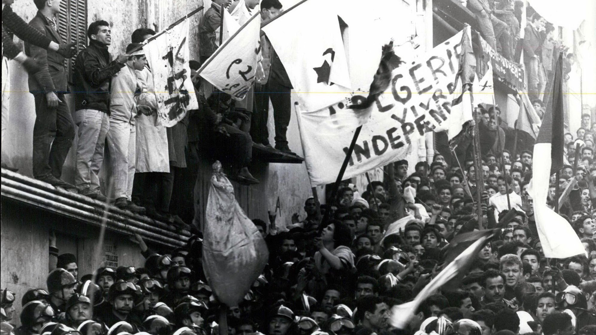L'image montre une grande foule rassemblée, avec des individus brandissant des drapeaux et des bannières. L'atmosphère semble tendue et chargeée d'émotion, probablement liée à un événement historique. Des personnes se tiennent sur un bâtiment, tandis que d'autres sont regroupées en bas, certaines portant des uniformes, ce qui pourrait indiquer une présence policière ou militaire. Le message sur les bannières évoque des thèmes d'indépendance, suggérant un contexte de luttes sociales ou politiques.