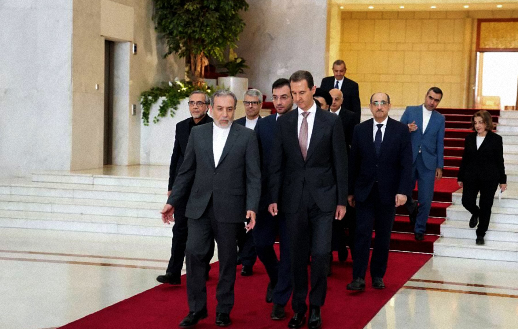 L'image montre un groupe de personnes marchant sur un tapis rouge dans un hall d'entrée élégant. À l'avant, deux hommes semblent en conversation, tandis que d'autres personnes les suivent, allant d'une manière formelle. L'environnement est spacieux avec des murs clairs et une décoration soignée, indiquant un lieu officiel ou diplomatique.