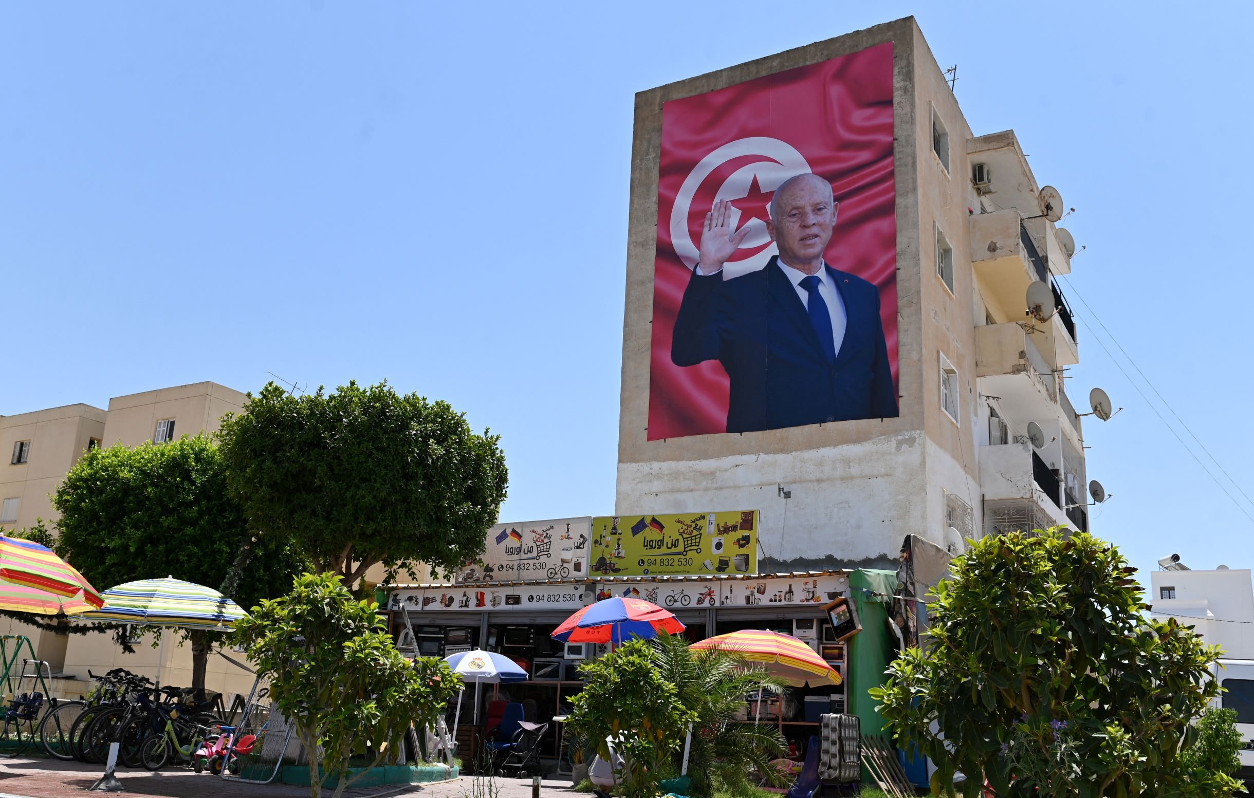 L'image montre un bâtiment avec une grande fresque représentant un homme, probablement un dirigeant, qui fait un geste de salutation. En arrière-plan, on peut voir un drapeau tunisien. Au premier plan, il y a des parasols colorés et des arbres, ainsi qu'une scène de vie qui pourrait inclure des personnes assises ou se déplaçant. Le ciel est clair et ensoleillé, ce qui donne une atmosphère lumineuse à la scène.