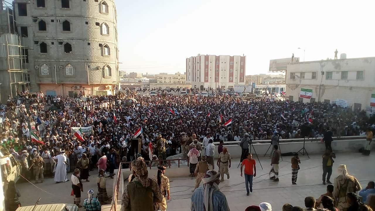 L'image montre une grande foule rassemblée lors d'une manifestation ou d'un rassemblement public. Les participants tiennent des banderoles et des drapeaux, indiquant un fort engagement politique ou social. En arrière-plan, on peut voir des bâtiments en construction ou rénovés, ainsi qu'une architecture variée. La scène semble se dérouler en plein air, probablement durant l'après-midi ou le début de la soirée, avec une lumière naturelle éclatante. L'atmosphère est dynamique, avec beaucoup de gens engagés dans une cause commune.