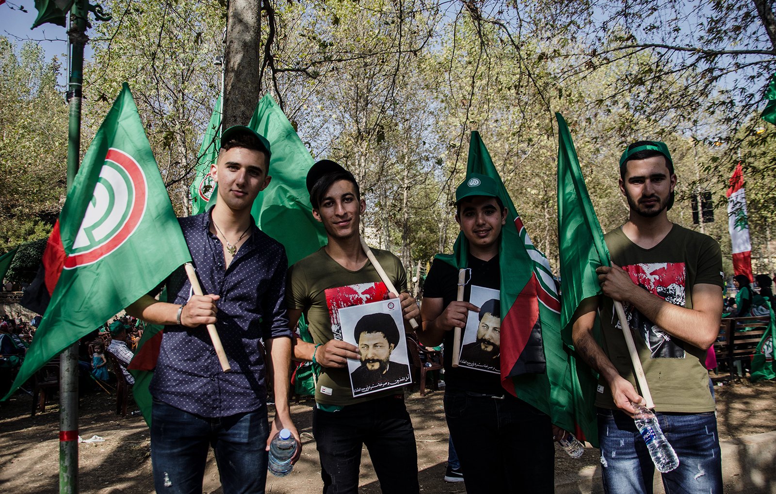 L'image montre un groupe de quatre jeunes hommes rassemblés lors d'un événement, probablement une manifestation ou une célébration. Ils tiennent des drapeaux verts, qui semblent avoir un symbole spécifique. Chacun d'eux porte des affiches représentant des figures, et ils affichent des expressions de fierté ou de détermination. L'arrière-plan est verdoyant, évoquant un cadre festif en plein air.