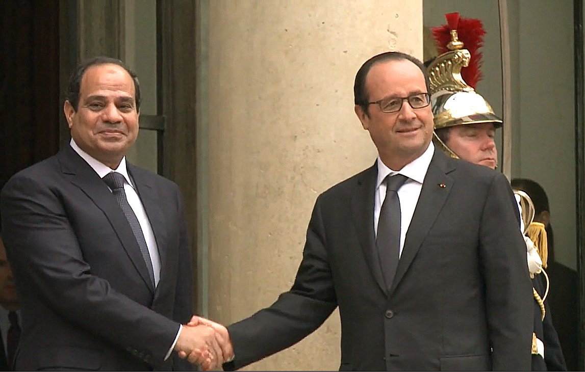 L'image montre deux hommes en costume, serrant la main l'un de l'autre. L'un est à gauche et semble avoir une expression amicale, tandis que l'autre se trouve à droite, affichant également un sourire. En arrière-plan, on aperçoit un soldat en uniforme. L'environnement semble être formel, probablement devant un bâtiment gouvernemental.