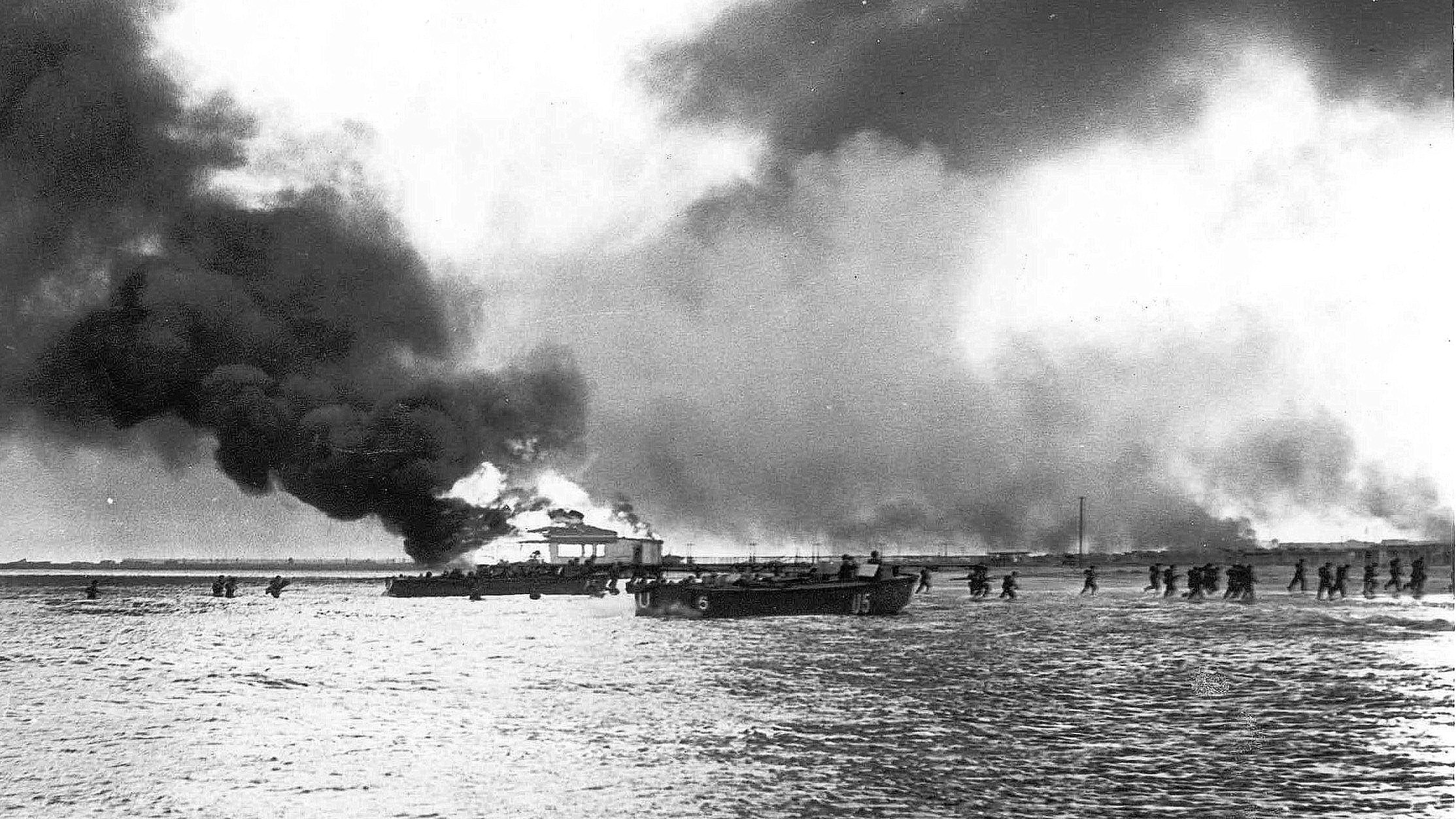 L'image montre une scène sombre et dramatique, probablement liée à un événement de guerre ou de désastre. On peut apercevoir une grande quantité de fumée noire s'élevant dans le ciel, suggérant qu'un bâtiment ou un navire est en train de brûler. Au premier plan, il y a un bateau sur l'eau, et au loin, plusieurs personnes semblent marcher le long de la rive, potentiellement évacuant une zone touchée par le feu ou un conflit. L'atmosphère générale est sombre et chaotique, avec des éléments de destruction visibles.