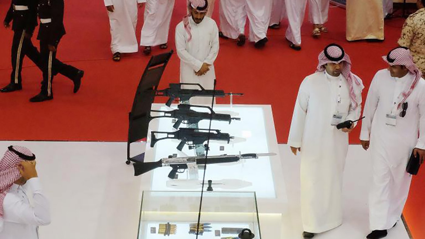 L'image montre un rassemblement où plusieurs personnes, probablement en tenue traditionnelle, se déplacent dans un espace décoré. Au centre, il y a une vitrine en verre contenant diverses armes, mettant en avant des fusils et d'autres équipements. L'ambiance semble formelle et le sol est recouvert d'un tapis rouge. Des membres de la sécurité ou des uniformes militaires peuvent être aperçus en arrière-plan, soulignant l'importance de l'événement.