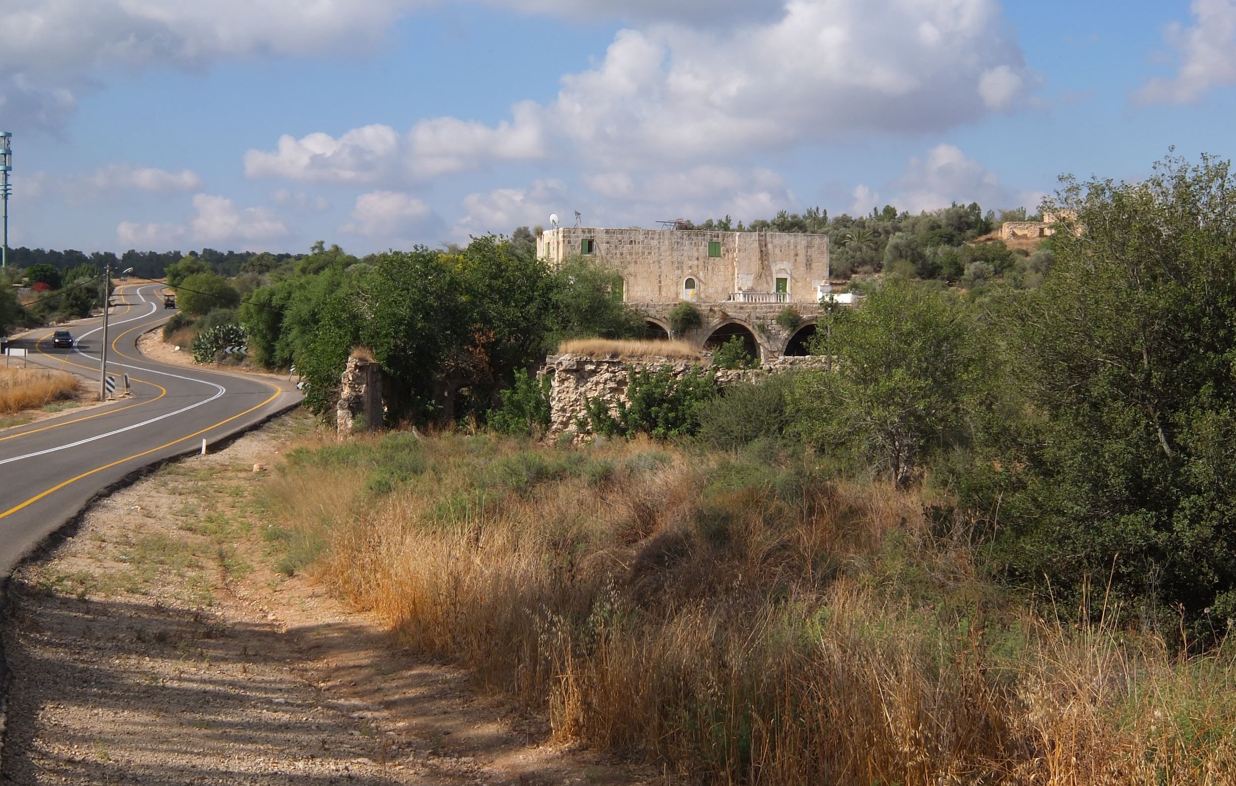 L'image montre un paysage rural avec une route sinueuse sur la gauche. À droite, il y a des herbes hautes et quelques arbres. Au fond, on aperçoit des ruines d'un bâtiment en pierre, probablement une ancienne construction, entourées de verdure. Le ciel est légèrement nuageux, créant une atmosphère paisible et naturelle.