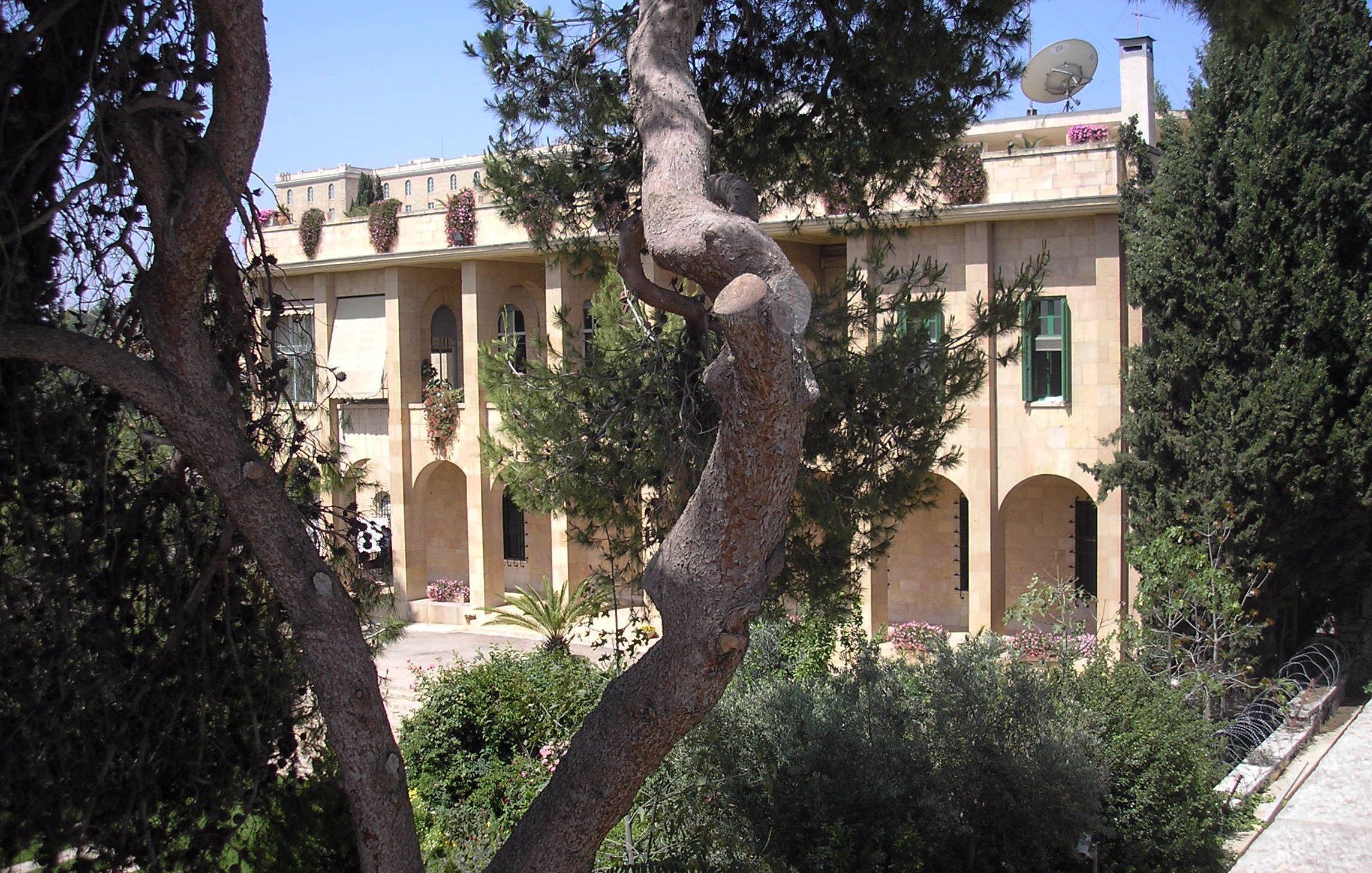 L'image montre un bâtiment en pierre, typique de l'architecture méditerranéenne, avec plusieurs arches et fenêtres. Il est entouré de verdure, avec des arbres et des plantes bien entretenues. Au fond, on remarque un satellite et un ciel bleu clair, ce qui suggère un climat ensoleillé. L'ensemble dégage une atmosphère paisible et accueillante.