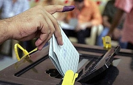 L'image montre une main avec un doigt marqué de violet, symbole d'un vote. La personne insère un bulletin de vote dans une urne électorale. En arrière-plan, on peut voir d'autres individus, probablement en train d'attendre ou de voter eux aussi. L'atmosphère semble sérieuse et engageante, illustrant un moment clé du processus démocratique.