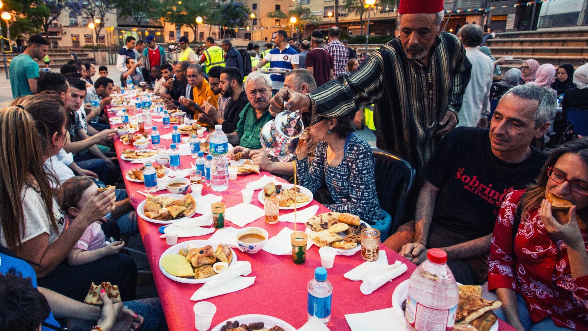 L'image montre une grande table dressée en plein air, où des personnes diverses sont réunies autour d'un repas convivial. Les convives, de différents âges, sont assis sur des chaises en plastique et profitent de divers plats, avec des assiettes remplies de nourriture. On peut voir des bouteilles d'eau sur la table et des verres. L'ambiance semble festive et chaleureuse, marquée par des sourires et des échanges entre les participants. En arrière-plan, on aperçoit d'autres personnes et des éléments de l'environnement urbain, indiquant une atmosphère de communauté.