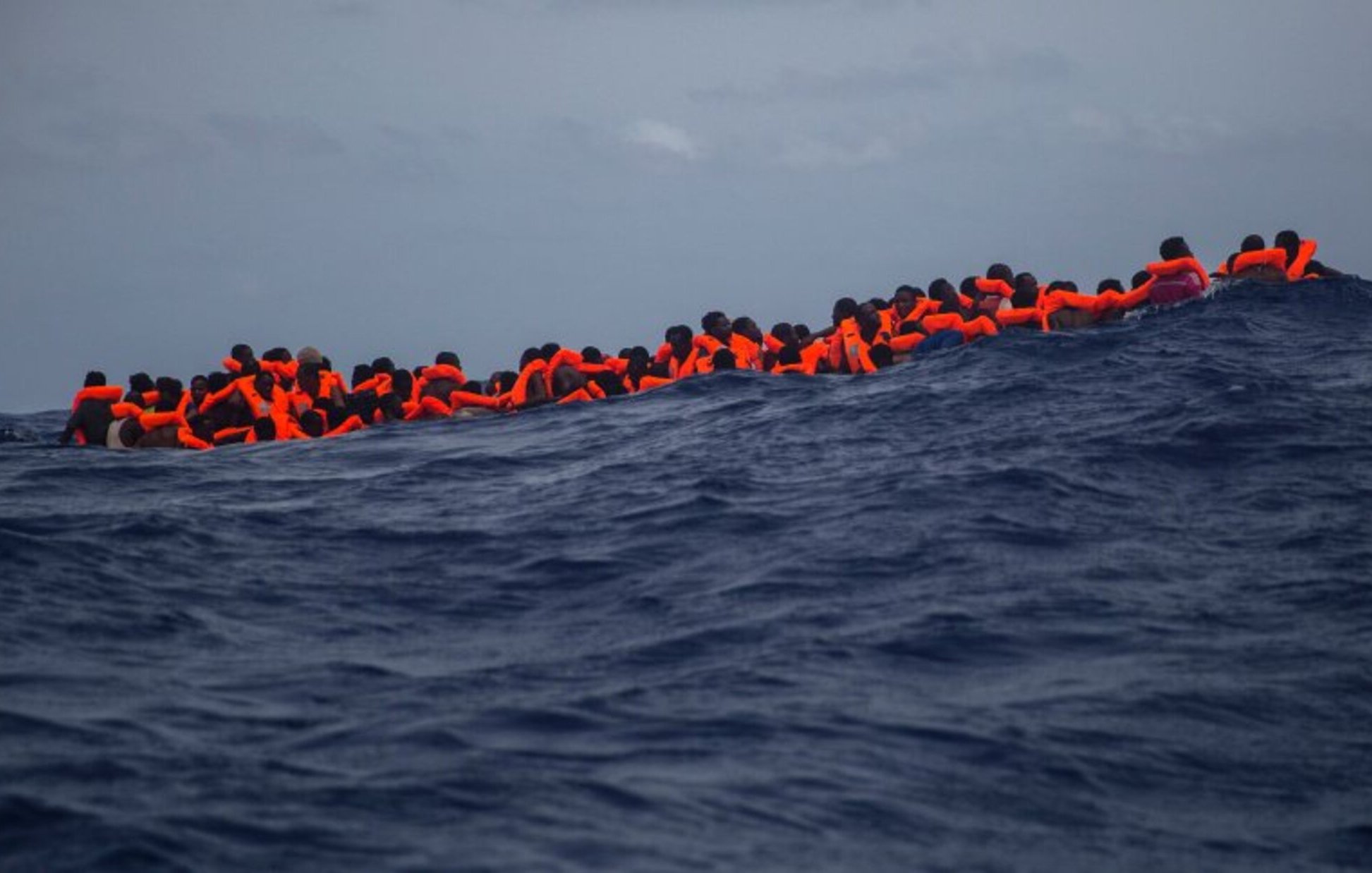 L'image montre un groupe de personnes en mer, portant des gilets de sauvetage orange. Elles semblent rassemblées sur une petite embarcation, luttant contre des vagues dans une étendue d'eau bleue profonde. Le ciel est nuageux, ce qui accentue la gravité de la situation maritime. Cette scène évoque des thèmes de migration, de survie et de danger en mer.