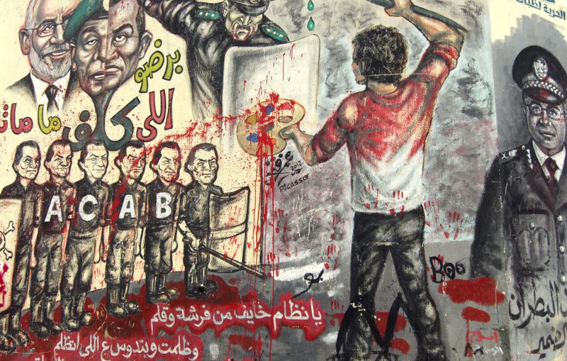 L'image montre un mural coloré, rempli de graffitis et d'illustrations. On y voit une scène dynamique où une personne, de dos, est en train de peindre avec un pinceau, probablement en train d'exprimer un message de frustration ou de résistance. Autour de cette figure, il y a diverses représentations de policiers et de figures autoritaires, certains avec des visages mécontents, indiquant une atmosphère de contestation. Le mur est éclaboussé de peinture rouge, ce qui pourrait symboliser la violence ou le sang, renforçant le message de lutte contre la répression. Les lettres "ACAB" sont visibles, ce qui fait référence à une phrase souvent associée à la critique des forces de police. L'ensemble de la composition semble porter un message fort sur l'opposition à l'autorité.