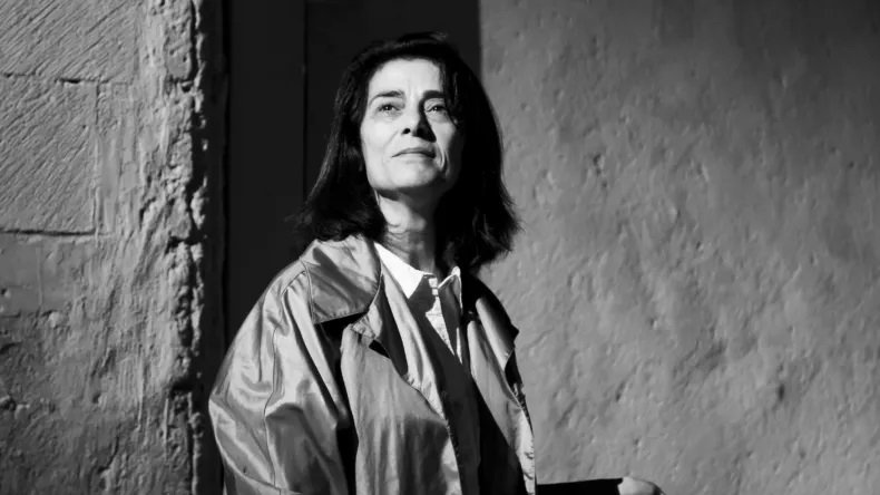 L'image présente une femme qui se tient debout, regardant légèrement vers le haut. Elle a les cheveux longs et foncés, et porte un manteau clair. La lumière joue sur son visage, créant un contraste fort avec l'arrière-plan, qui semble être texturé et neutre. L'ensemble de l'image est en noir et blanc, ce qui accentue l'intensité de l'expression de son visage et la profondeur de la scène.