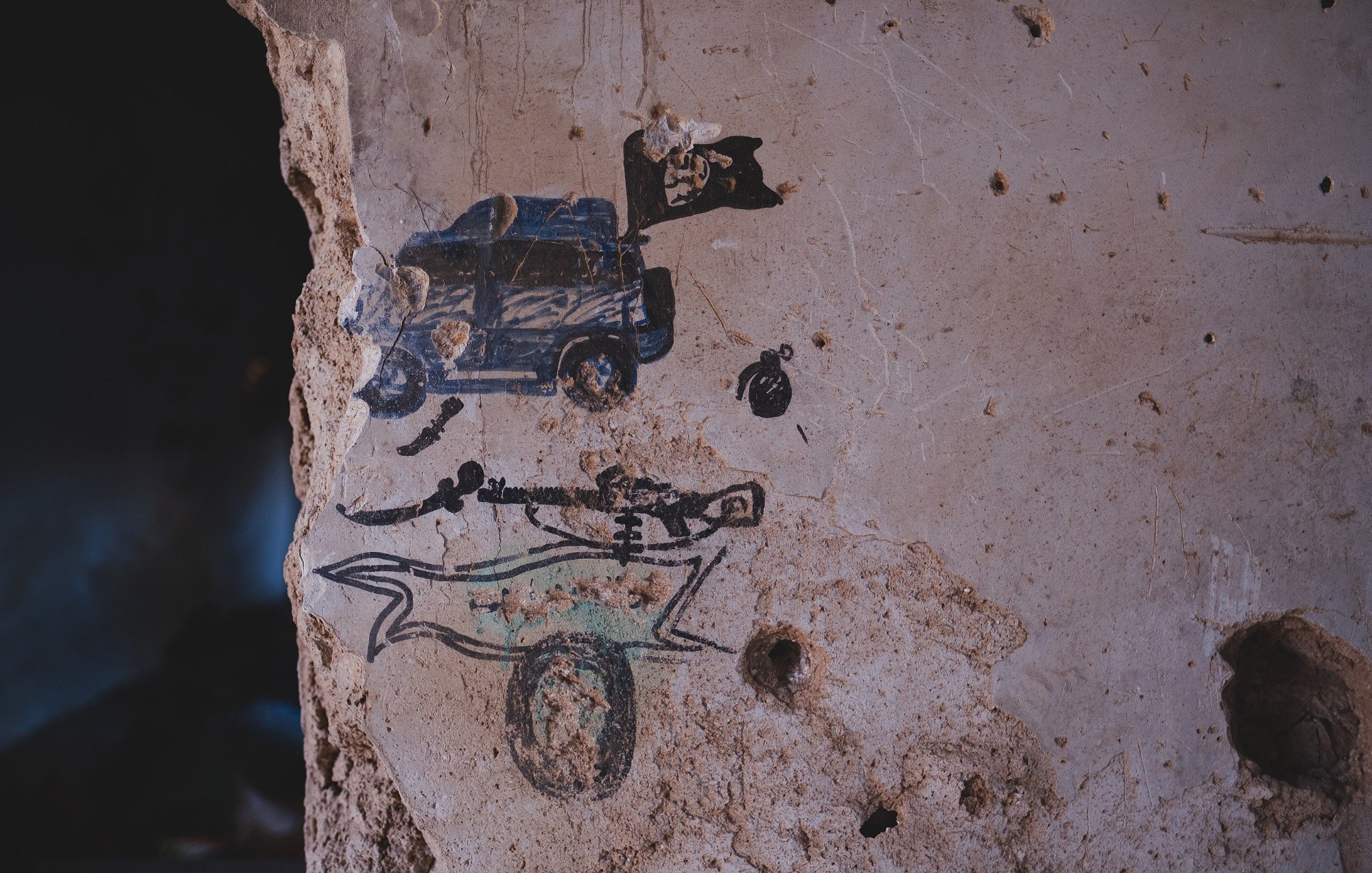 L'image montre un mur en béton décrépit où des graffitis sont visibles. On peut y voir une représentation d'un véhicule, probablement une voiture de police, accompagnée d'un drapeau. En dessous, il y a un dessin stylisé d'un squelette, surmonté d'une forme qui pourrait évoquer un traîneau ou un chariot. Le mur lui-même présente des traces d'usure et des fissures, ajoutant une dimension d'abandon à la scène.