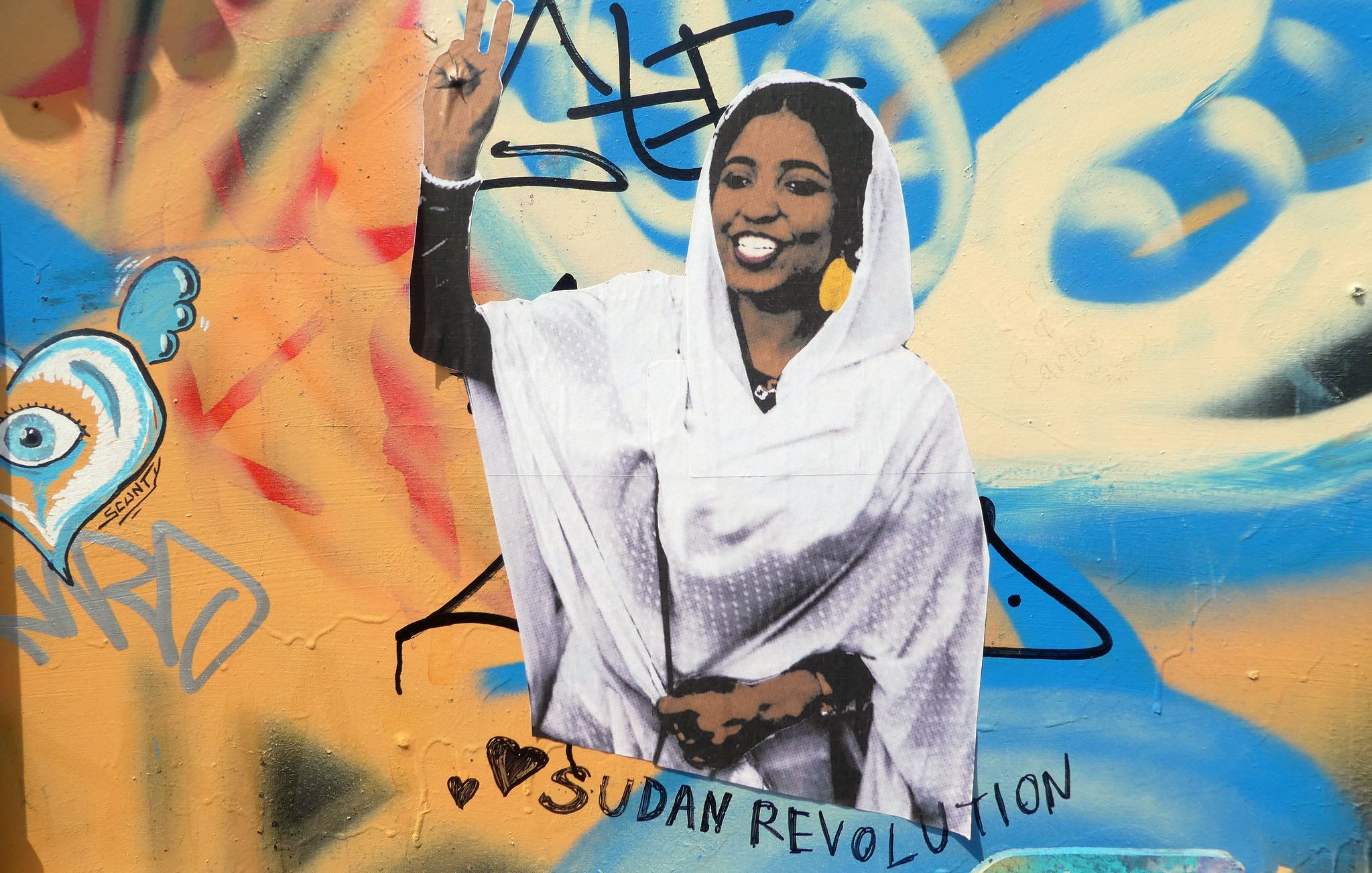 L'image montre une peinture murale colorée sur un mur. Au centre, il y a une représentation d'une femme vêtue d'une robe blanche, levant la main en signe de victoire avec deux doigts. Elle affiche un large sourire, symbolisant la positivité et la résistance. Autour d'elle, il y a des graffitis multicolores, et l'inscription "Sudan Revolution" est bien visible, ce qui indique un lien avec le mouvement révolutionnaire au Soudan. Le tout dégage une ambiance de soutien et de solidarité.