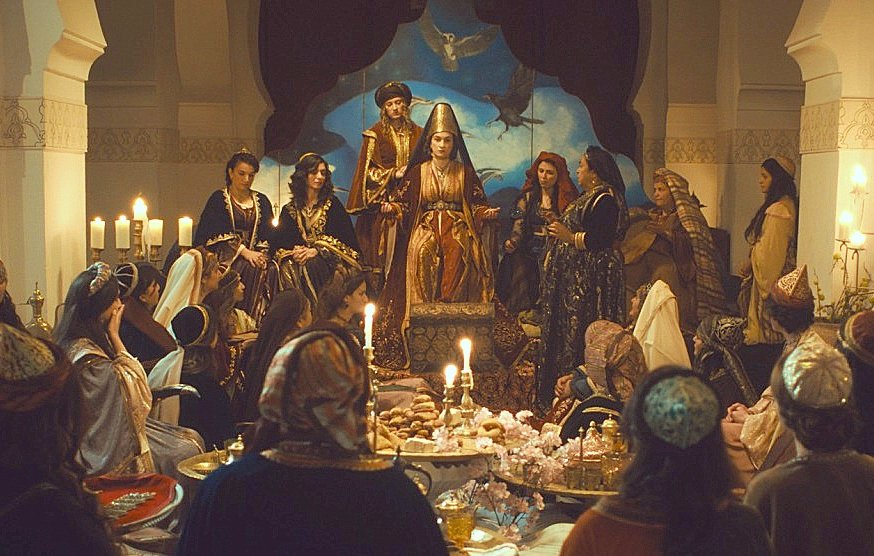 L'image montre une scène richement décorée, probablement d'inspiration historique ou fantastique. Au centre, une figure majestueuse est assise sur un trône, entourée de femmes habillées de vêtements ornés. L'ambiance est chaleureuse et conviviale, éclairée par des chandeliers et ambiance de festin, avec des plats disposés sur de grandes tables. Le fond est orné de motifs artistiques, évoquant une atmosphère de cour royale ou de réunion importante. Les expressions des personnages laissent entrevoir des interactions vraisemblablement animées.