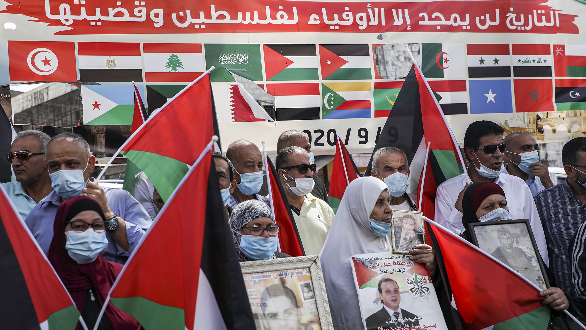 L'image montre un groupe de personnes rassemblées, portant des masques et tenant des drapeaux. Ils semblent manifester pour soutenir la Palestine, avec des affiches et des portraits en mains. En arrière-plan, on peut voir une grande bannière ornée de multiples drapeaux de différents pays. L'ambiance est engagée et solidaire, témoignant d'une mobilisation pour une cause politique.