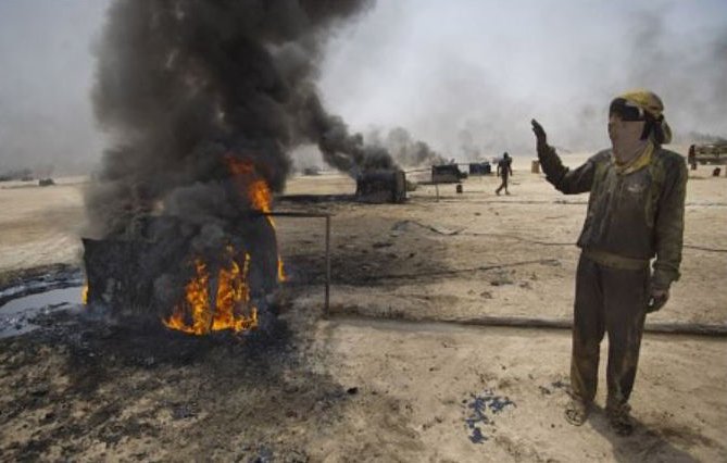L'image montre un homme dans un environnement industrialisé, à proximité de réservoirs en feu. Des flammes et une épaisse fumée noire s'élèvent de plusieurs conteneurs. L'homme semble faire un geste d'alerte ou de signalisation, et il porte des vêtements de travail, suggérant qu'il se trouve dans une zone à risque, possiblement liée à l'exploitation pétrolière. En arrière-plan, d'autres personnes sont visibles, mais leur activité n'est pas clairement définie. L'atmosphère de la scène est sombre et montre les dangers liés aux incendies industriels.