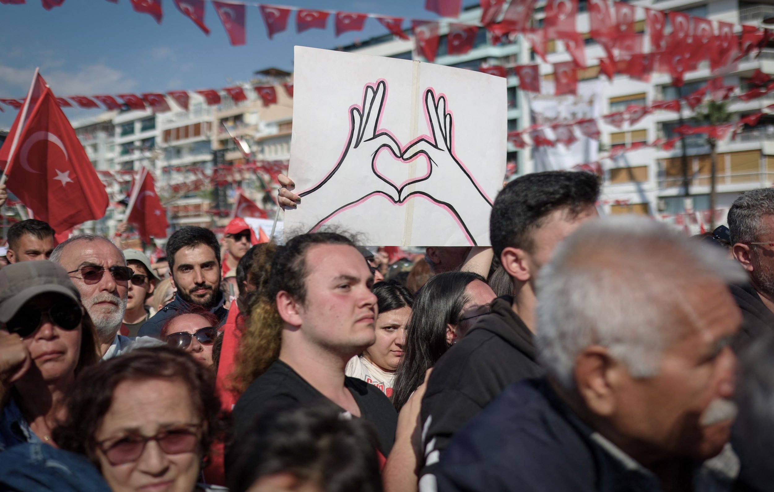 L'image montre une grande foule rassemblée lors d'un événement en plein air. Au centre, une personne tient une pancarte avec un dessin de deux mains formant un cœur. La majorité des personnes présentes portent des vêtements aux couleurs nationales, notamment du rouge et du blanc, et certains affichent des drapeaux turcs. L'ambiance semble festive et engagée, témoignant d'une manifestation ou d'une célébration collective.