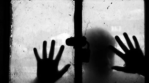 L'image montre une fenêtre sale avec un verre embué. Deux mains sont posées sur le verre, comme si quelqu'un tentait d'atteindre l'extérieur. La silhouette d'une personne est visible derrière le verre, ajoutant une atmosphère mystérieuse et mélancolique à la scène. L'utilisation du noir et blanc accentue le contraste et l'émotion de l'image, créant une sensation d'isolement et de confinement.