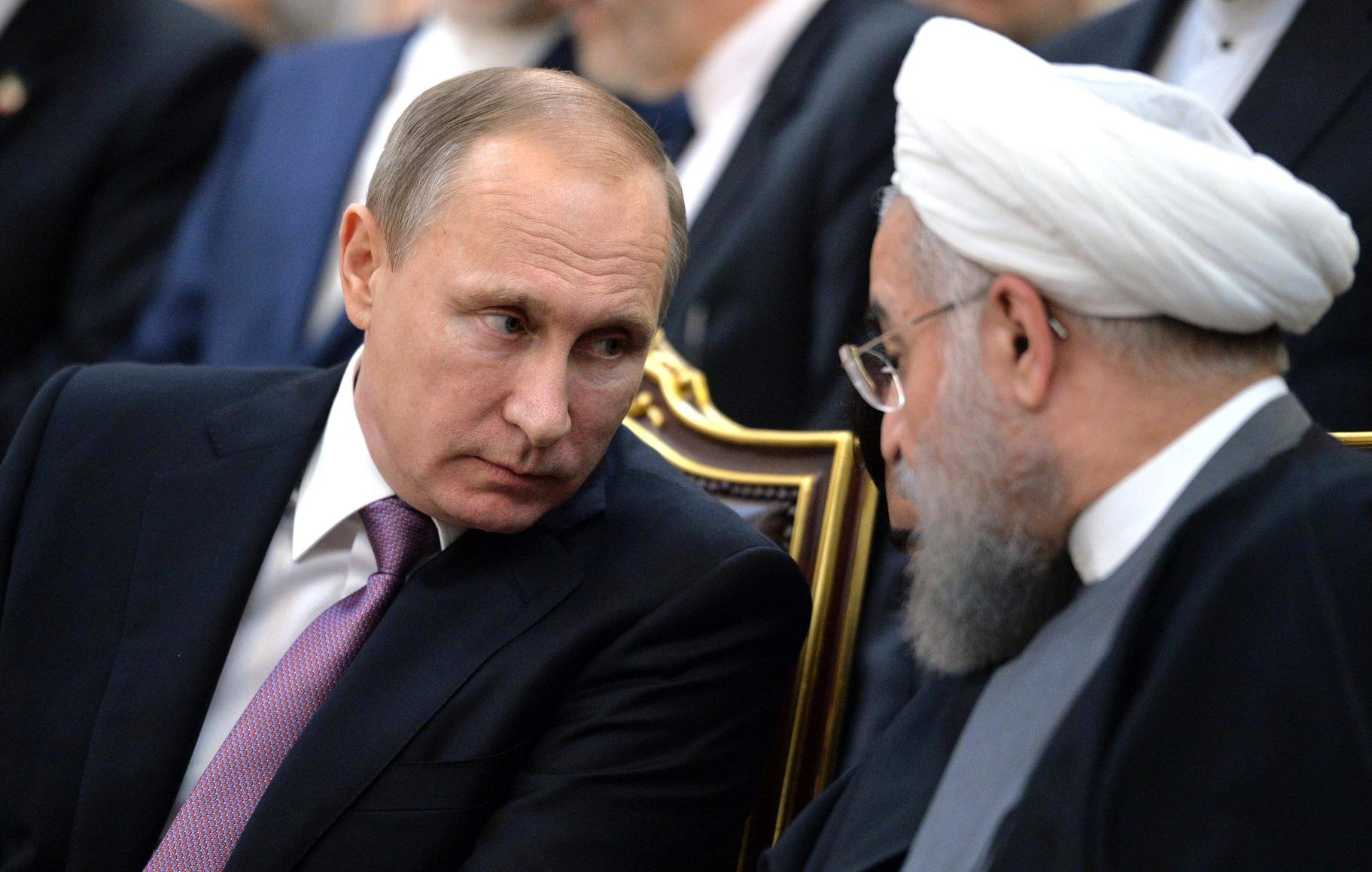 L'image montre deux hommes en train de discuter, assis côte à côte. L'un porte un costume noir avec une cravate et a un regard sérieux, tandis que l'autre est en tenue traditionnelle avec un turban et une barbe. Leurs expressions reflètent une conversation attentive. L'environnement est formel, suggérant un contexte politique ou diplomatique.