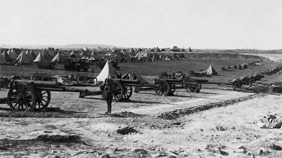 L'image montre un camp militaire avec plusieurs canons en premier plan. À l'arrière-plan, on distingue des tentes disposées de manière ordonnée sur un terrain dégagé. Le paysage semble vaste, avec des collines ou des montagnes au loin. L'atmosphère est historique, évoquant une période de guerre ou de mobilisation des troupes. Un soldat ou un homme se tient près des canons, donnant une échelle à la scène.