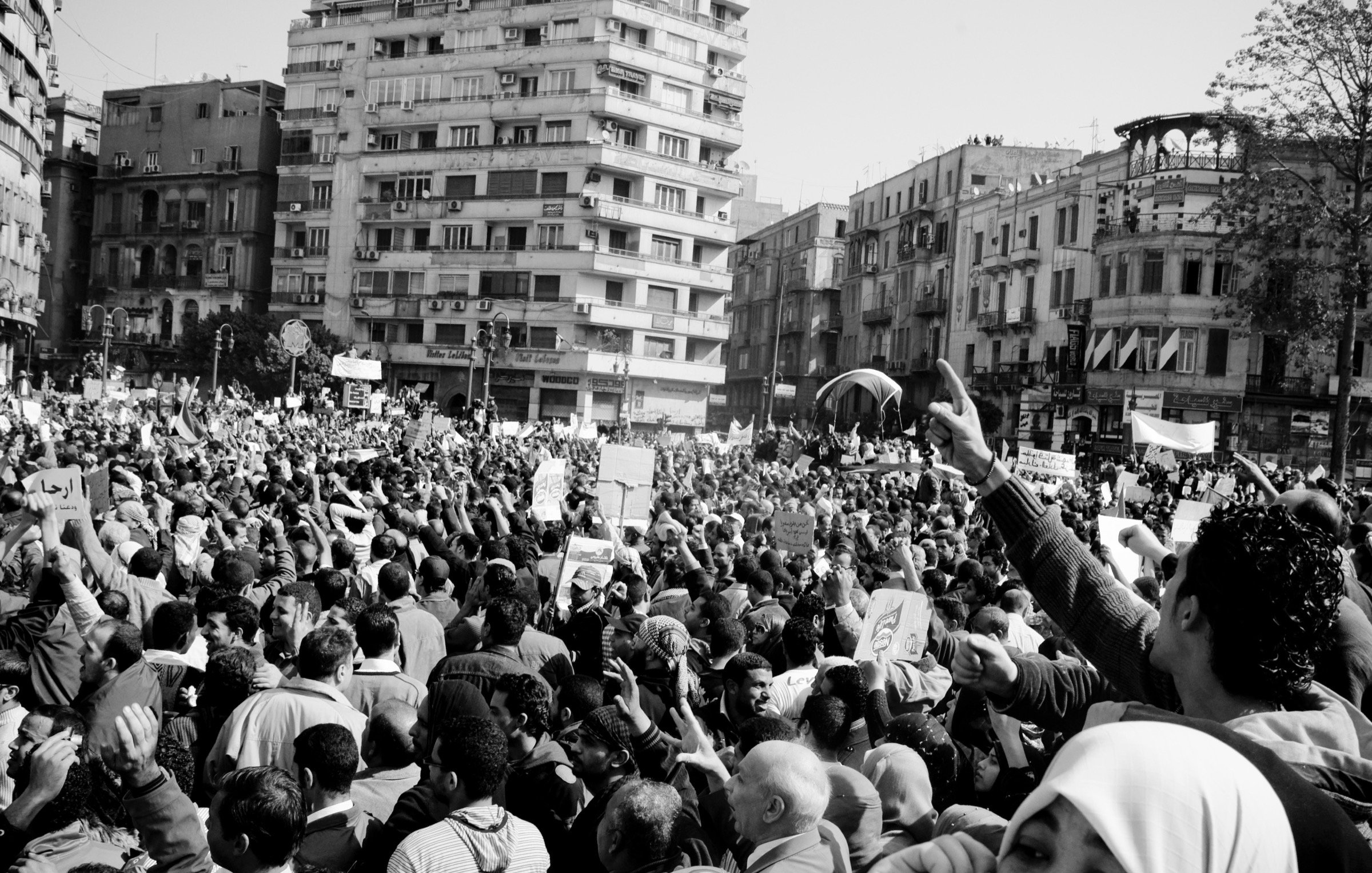 L'image montre une grande foule rassemblée dans une ville, probablement lors d'une manifestation ou d'une protestation. Les participants brandissent des pancartes et semblent exprimer des opinions fortes. L'atmosphère est dynamique et engagée, avec beaucoup de personnes levées les bras, ce qui suggère une ambiance de mobilisation collective. Le noir et blanc de l'image accentue les émotions et l'intensité du moment. Les bâtiments environnants ajoutent un contexte urbain à cette scène de rassemblement.
