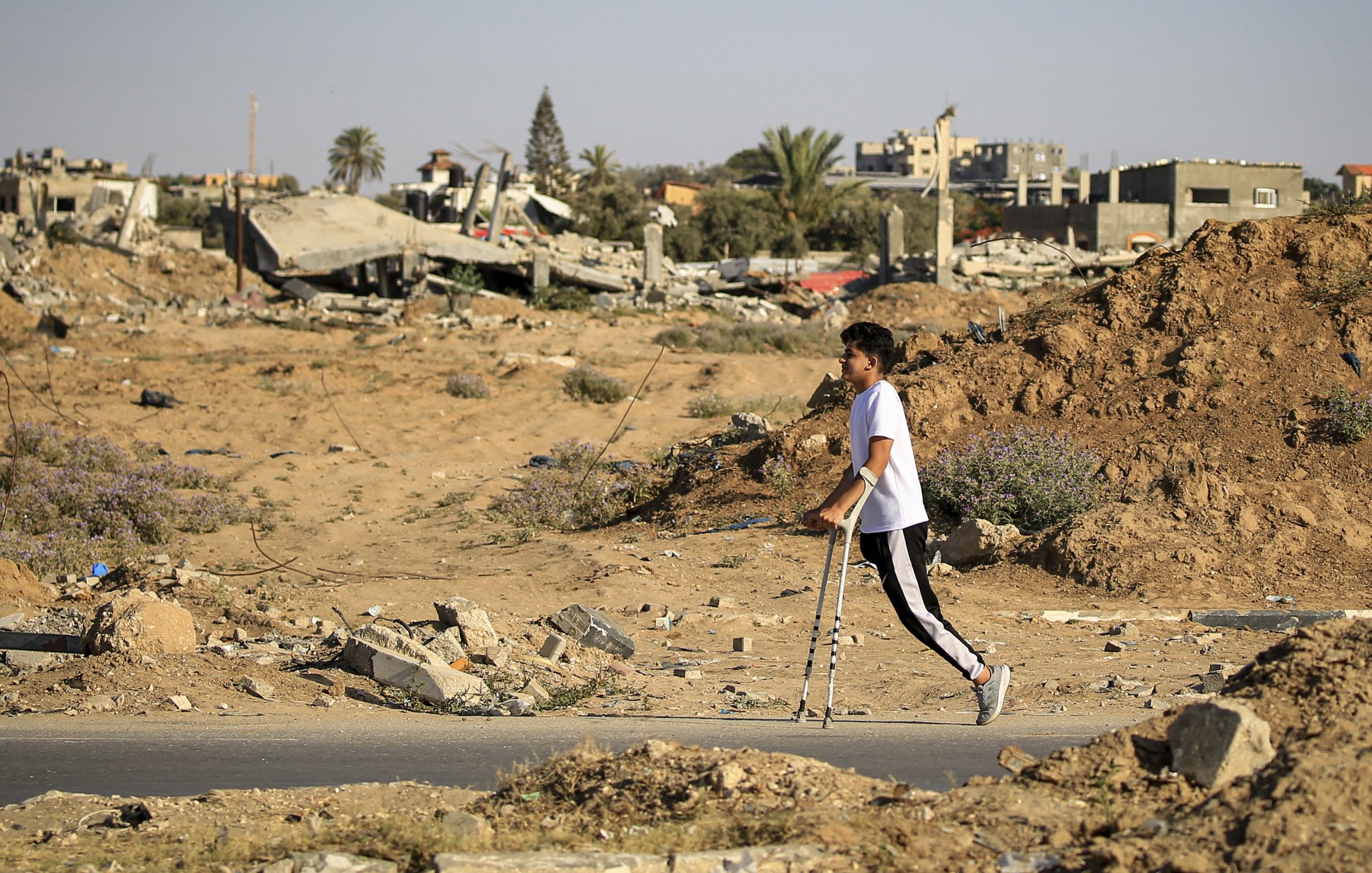 L'image montre un jeune garçon marchant avec des béquilles dans un paysage de destruction. En arrière-plan, on peut voir des bâtiments en ruines et des débris éparpillés, témoignant d'un environnement dévasté. Le sol est recouvert de terre et de petites herbes, créant un contraste avec les restes des structures. L'atmosphère semble sombre et tragique, soulignant la situation difficile dans laquelle se trouve l'enfant.