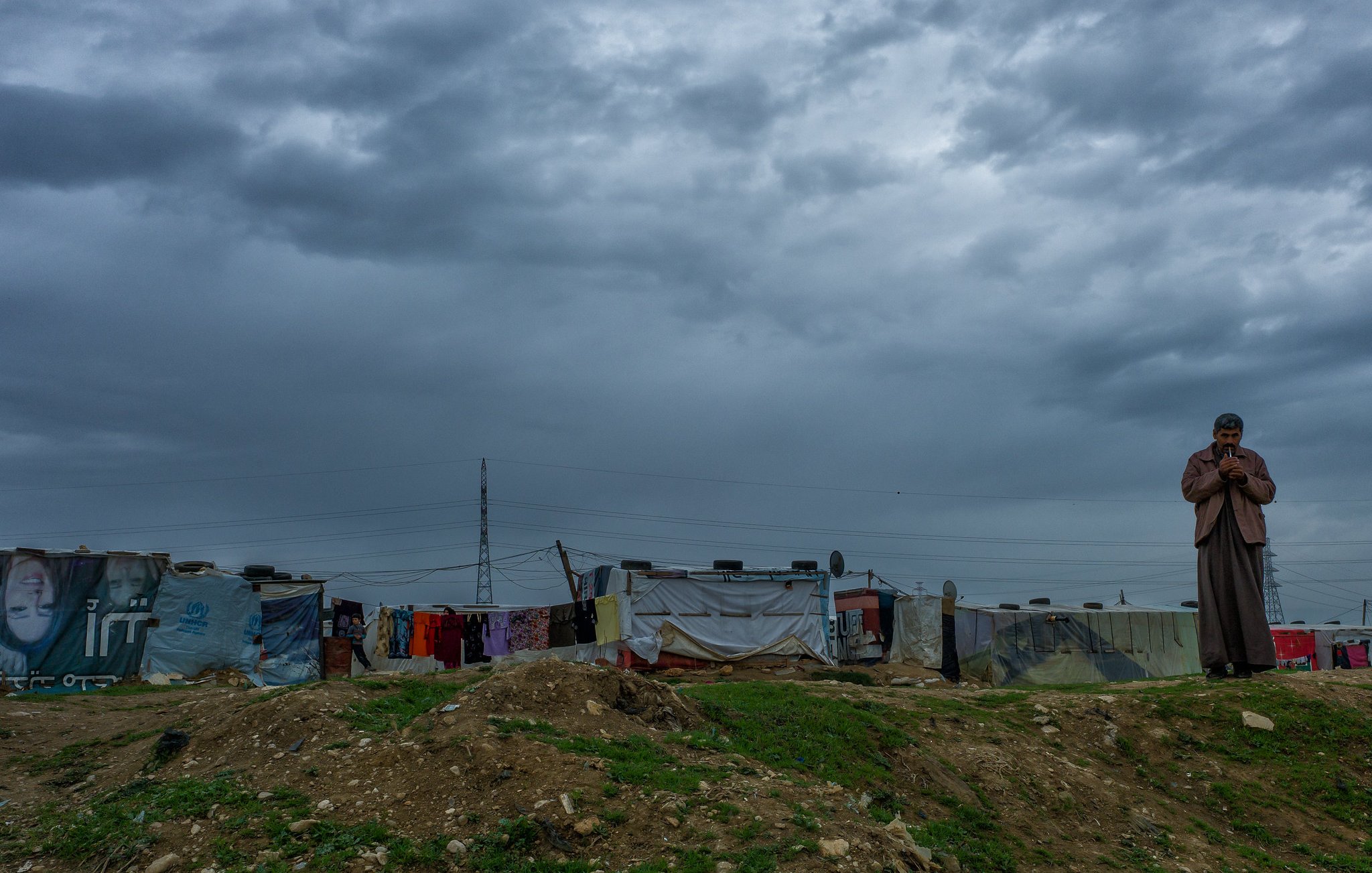 L'image montre une scène en extérieur sous un ciel nuageux. À droite, un homme se tient debout, apparemment pensif, avec une main près de son visage. En arrière-plan, on voit une rangée de tentes ou abris, probablement dans un camp, avec des murs en toile de différentes couleurs. Le sol est herbeux et légèrement surélevé. L'atmosphère est sombre en raison des nuages menaçants qui couvrent le ciel.