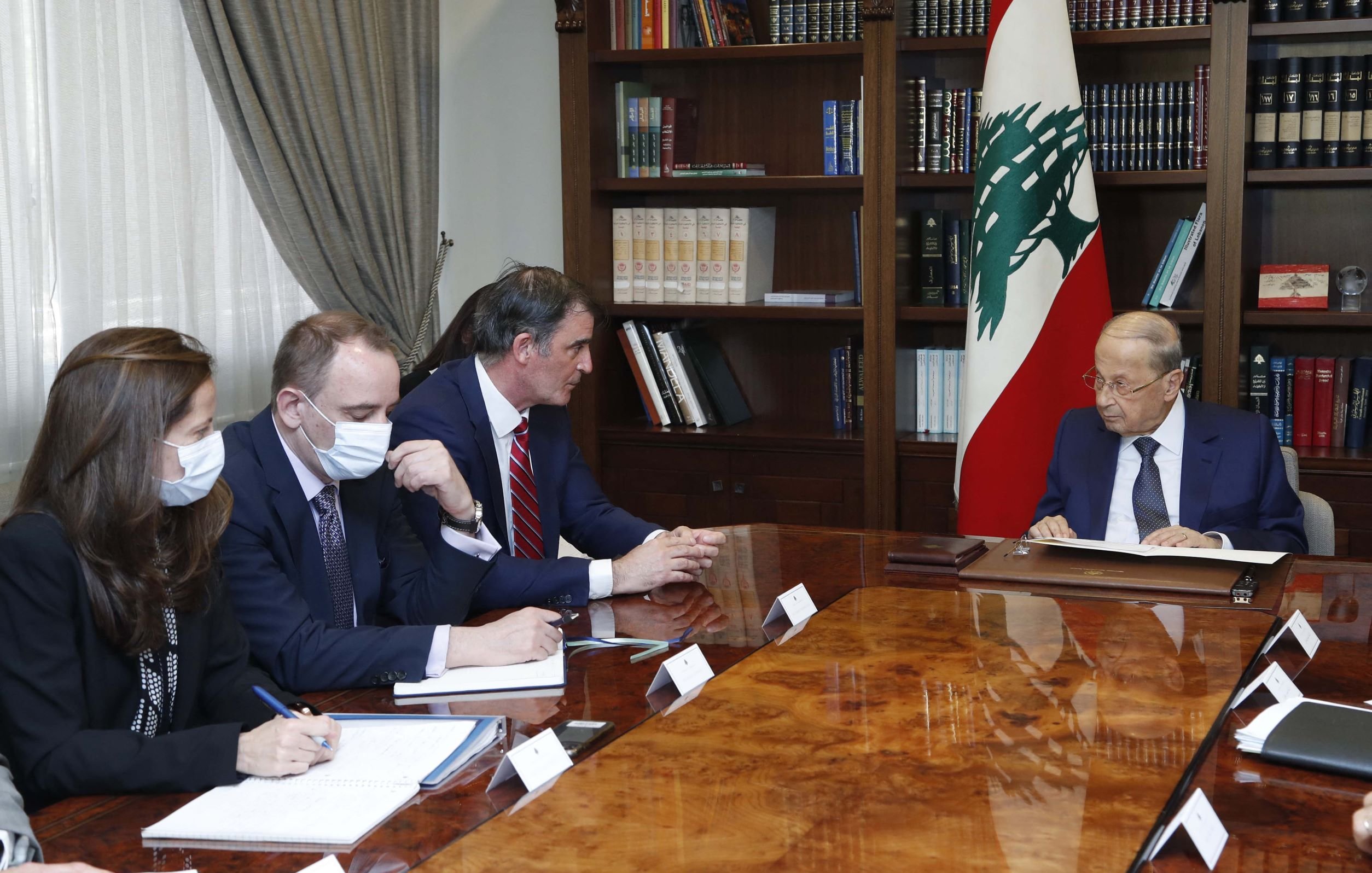 L'image montre une réunion dans un bureau. Un homme, probablement un leader politique, est assis à droite, face à une table en bois. Il semble concentré sur des documents. À sa gauche, il y a trois personnes, vraisemblablement des conseillers ou des diplomates, qui prennent des notes et écoutent attentivement. Tous portent des masques. En arrière-plan, on voit une bibliothèque remplie de livres et le drapeau du Liban. L'atmosphère semble formelle et sérieuse.