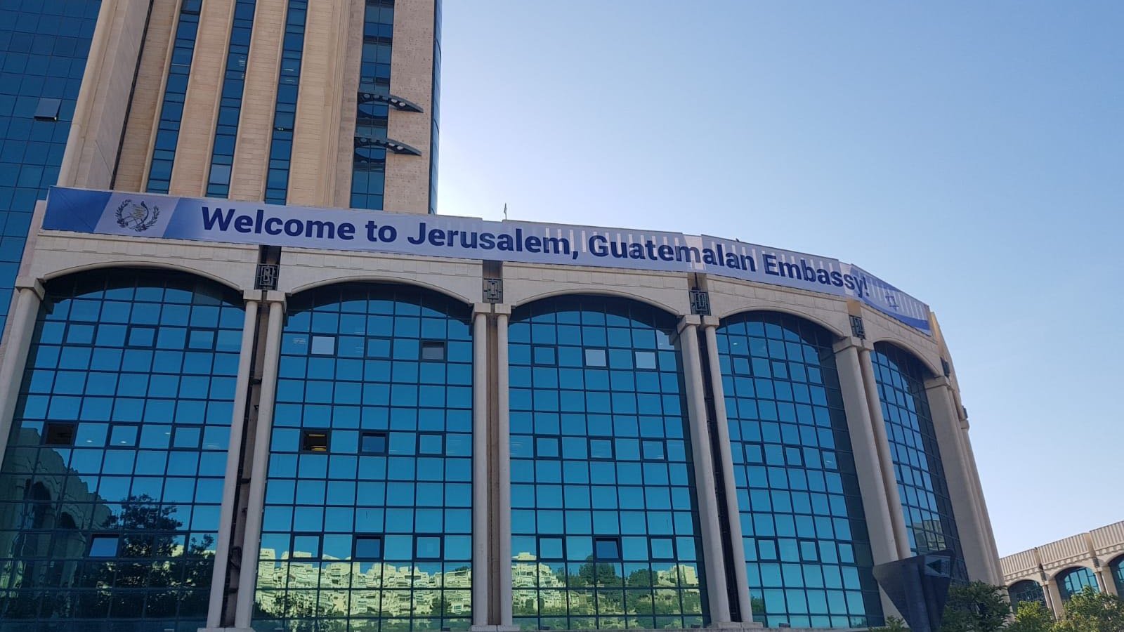 L'image montre un bâtiment moderne, probablement un immeuble administratif, avec une façade en verre réfléchissant. Au-dessus des fenêtres, une grande banderole affiche le message "Welcome to Jerusalem, Guatemalan Embassy". Le ciel est dégagé, suggérant une belle journée. Des arbres et des plantes ornent le devant du bâtiment, ajoutant une touche de verdure à l'environnement urbain.