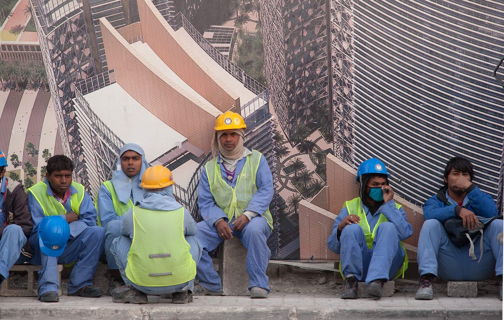 L'image montre un groupe de travailleurs assis sur un mur, probablement en train de faire une pause. Ils portent des vêtements de travail, notamment des gilets de sécurité et des casques de différentes couleurs. L'arrière-plan présente une grande affiche ou un panneau représentant un bâtiment moderne, suggérant qu'ils travaillent sur un site de construction. L'environnement semble urbain et dynamique.