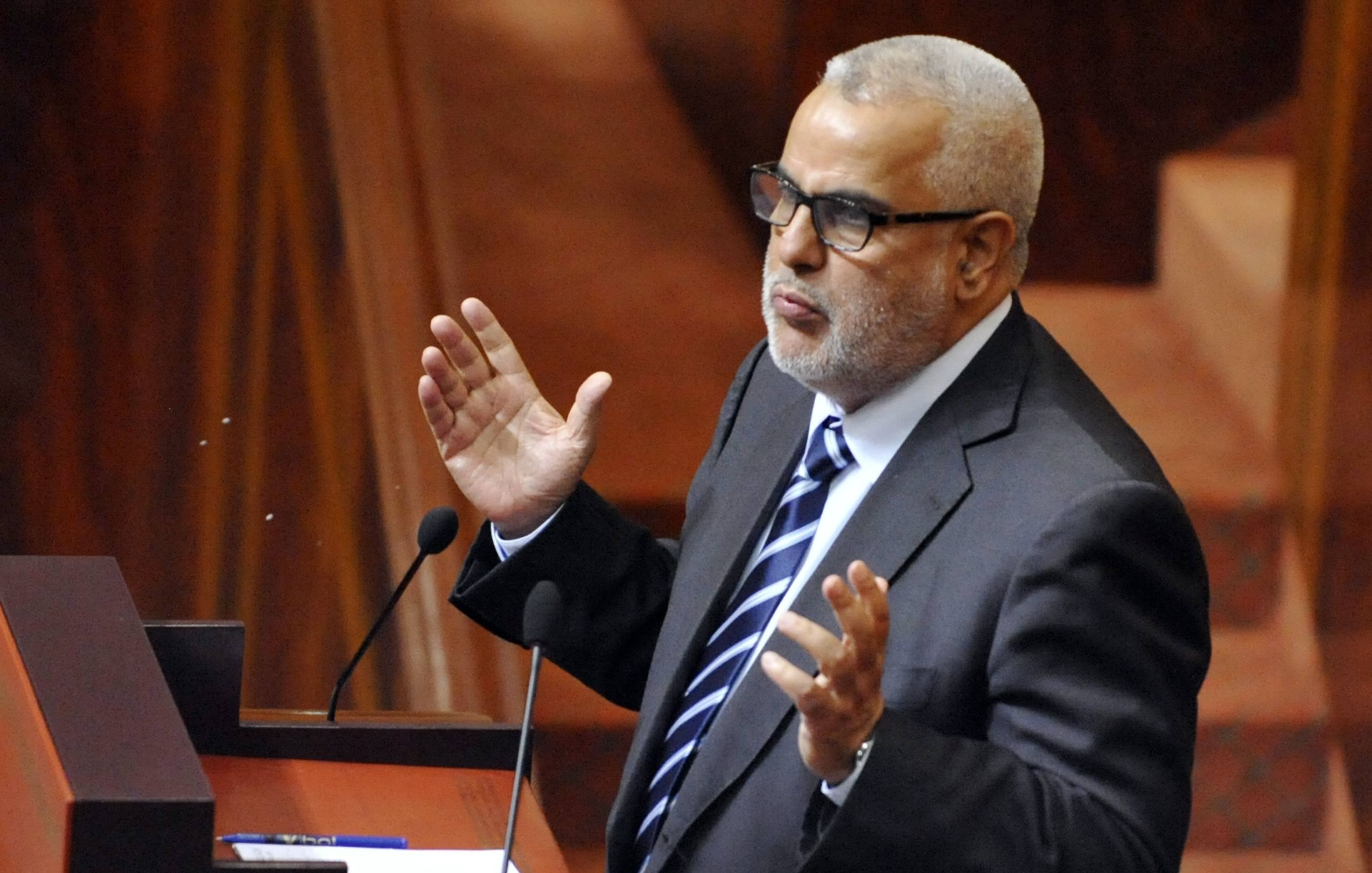 L'image montre un homme debout devant un pupitre, probablement en train de s'exprimer lors d'une réunion ou d'une séance législative. Il porte un costume sombre avec une chemise et une cravate à rayures. Son attitude et ses gestes suggèrent qu'il est engagé dans un discours ou une argumentation. L'environnement semble être celui d'une assemblée, avec des éléments de décor en bois et des sièges visibles en arrière-plan.