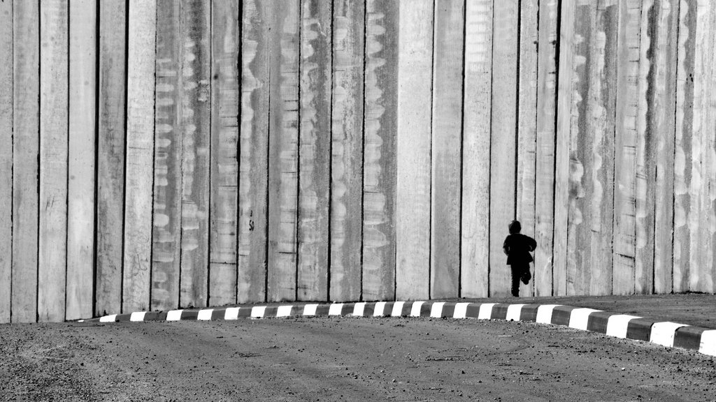 L'image montre un enfant courant le long d'un mur de béton. Le mur est haut et semble imposant, créant une ambiance de séparation ou d'isolement. Le contraste en noir et blanc accentue les textures du mur et la silhouette de l'enfant, qui semble minuscule par rapport à la structure massive. Le sol est désertique avec une route qui tourne légèrement, ajoutant à l'impression de solitude et de mouvement.