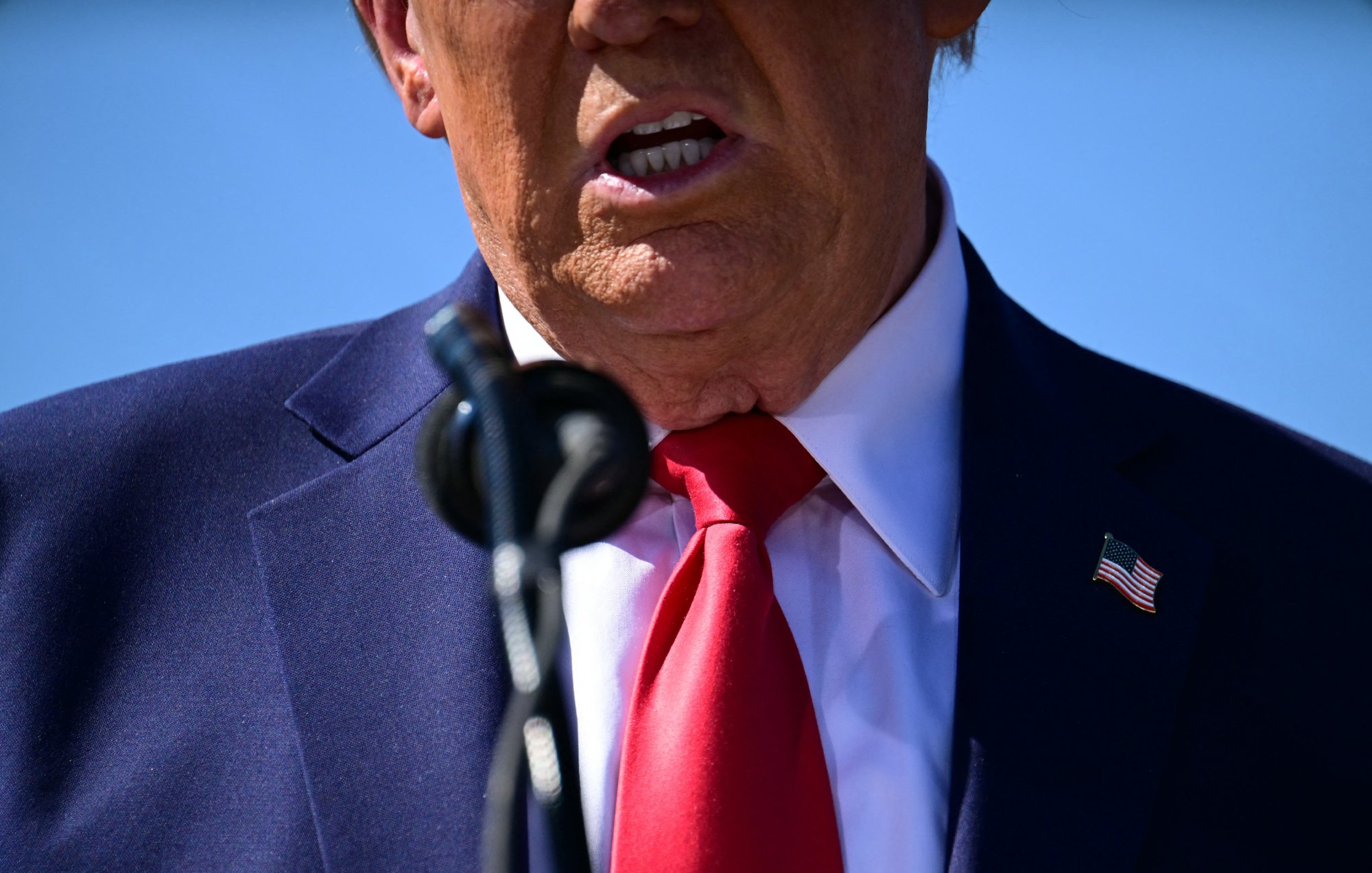 L'image montre principalement une partie supérieure du corps d'une personne, incluant sa bouche et son cou. La personne porte une chemise blanche sous un costume sombre, avec une cravate rouge. On peut également apercevoir un microphone devant elle. L'arrière-plan semble être un ciel bleu, suggérant un événement en extérieur.