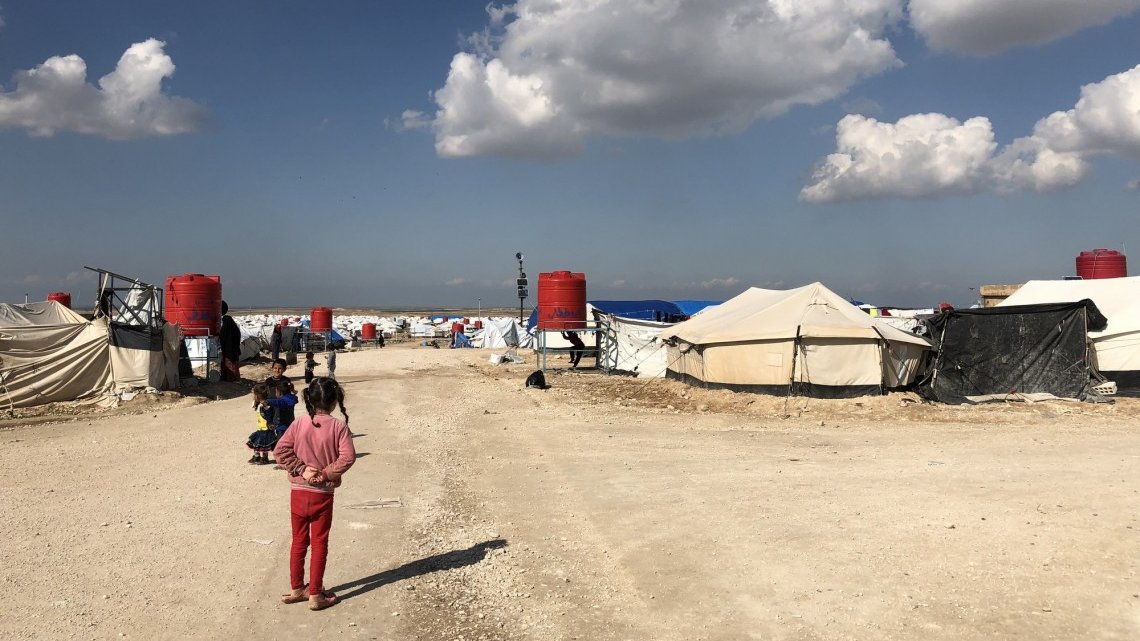 L'image montre un camp de réfugiés avec des tentes disposées de part et d'autre d'un chemin de terre. Au fond, on aperçoit des nuages blancs dans un ciel bleu. Des enfants sont présents, certains semblent jouer ou se déplacer dans le camp. Les abris sont principalement en toile, avec quelques réservoirs d'eau visibles. L'atmosphère paraît calme malgré les conditions de vie apparentes de ces lieux.